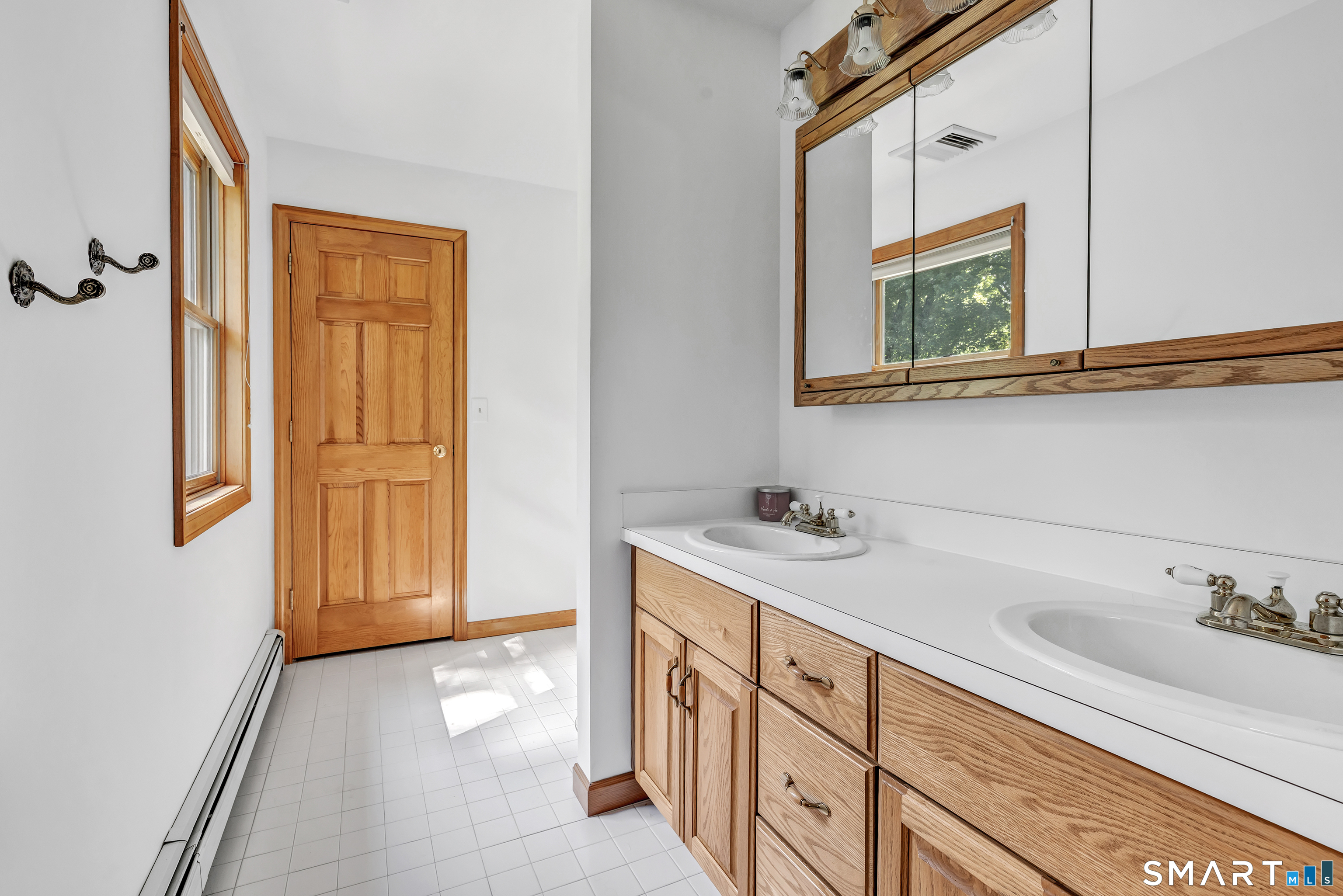 Bathroom with a double-sink vanity, light wood cabinetry, mirrored cabinet, and tiled floor.