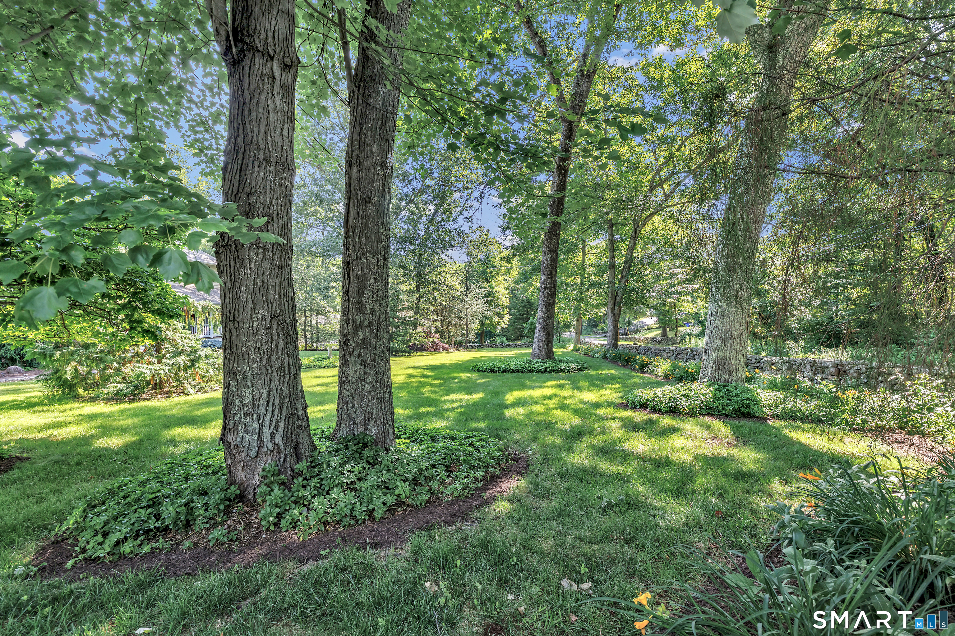 Landscaped yard with mature trees, a broad lawn, planted beds, and a low stone wall.