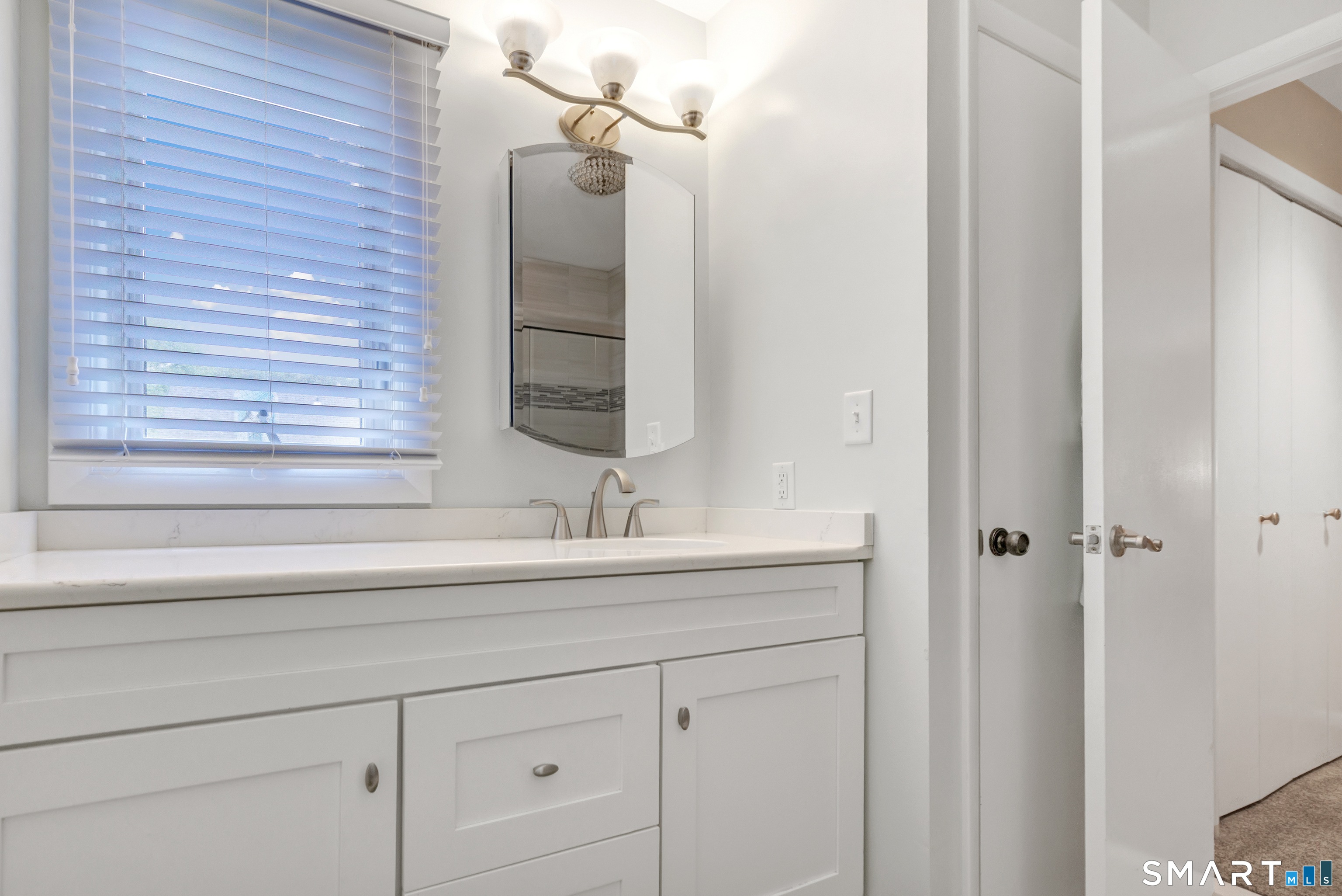 Bathroom vanity with white paneled cabinets, a mirrored medicine cabinet, and a window with blinds.
