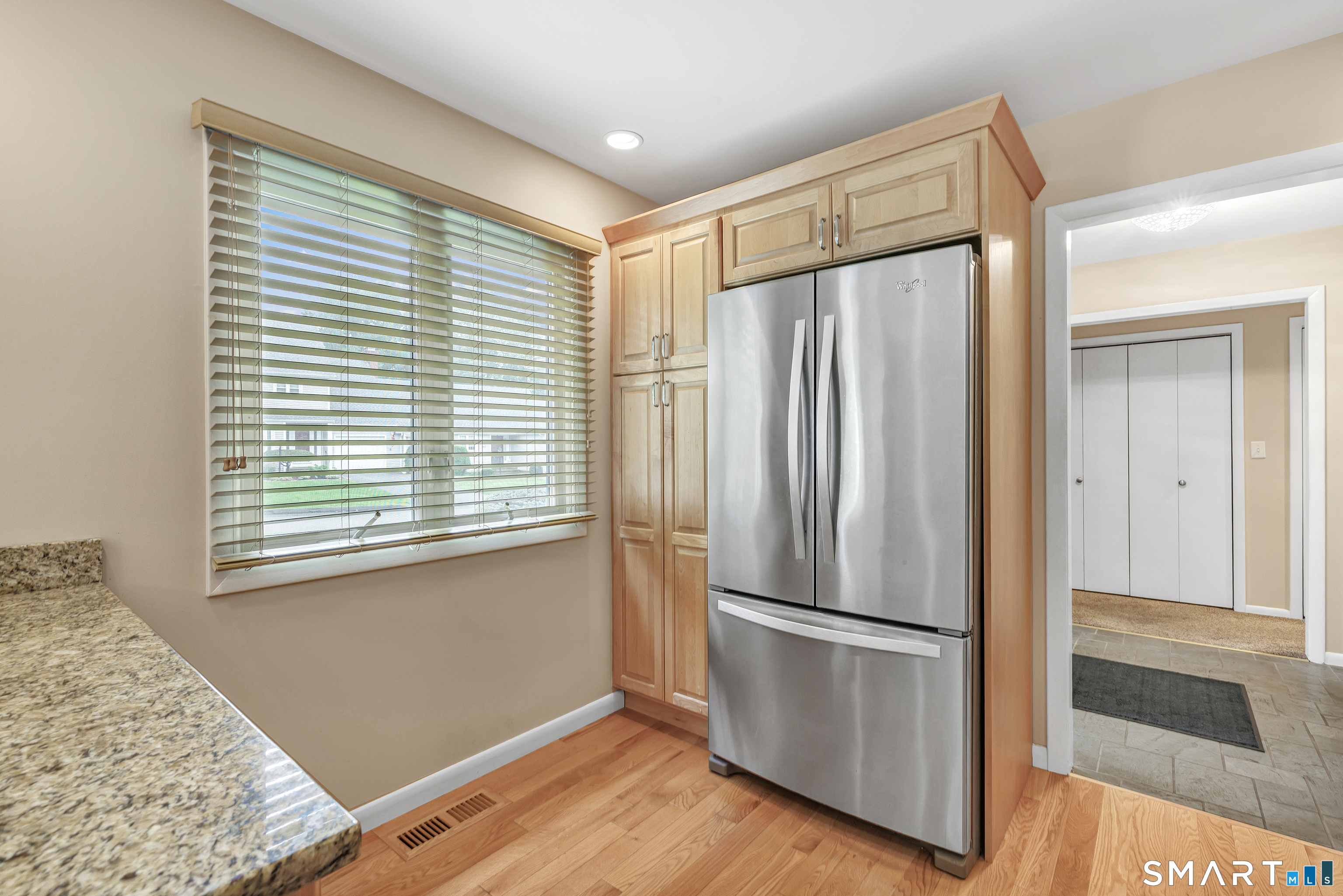 Kitchen with stainless steel refrigerator, light wood cabinetry, granite counter, and a large window with blinds.