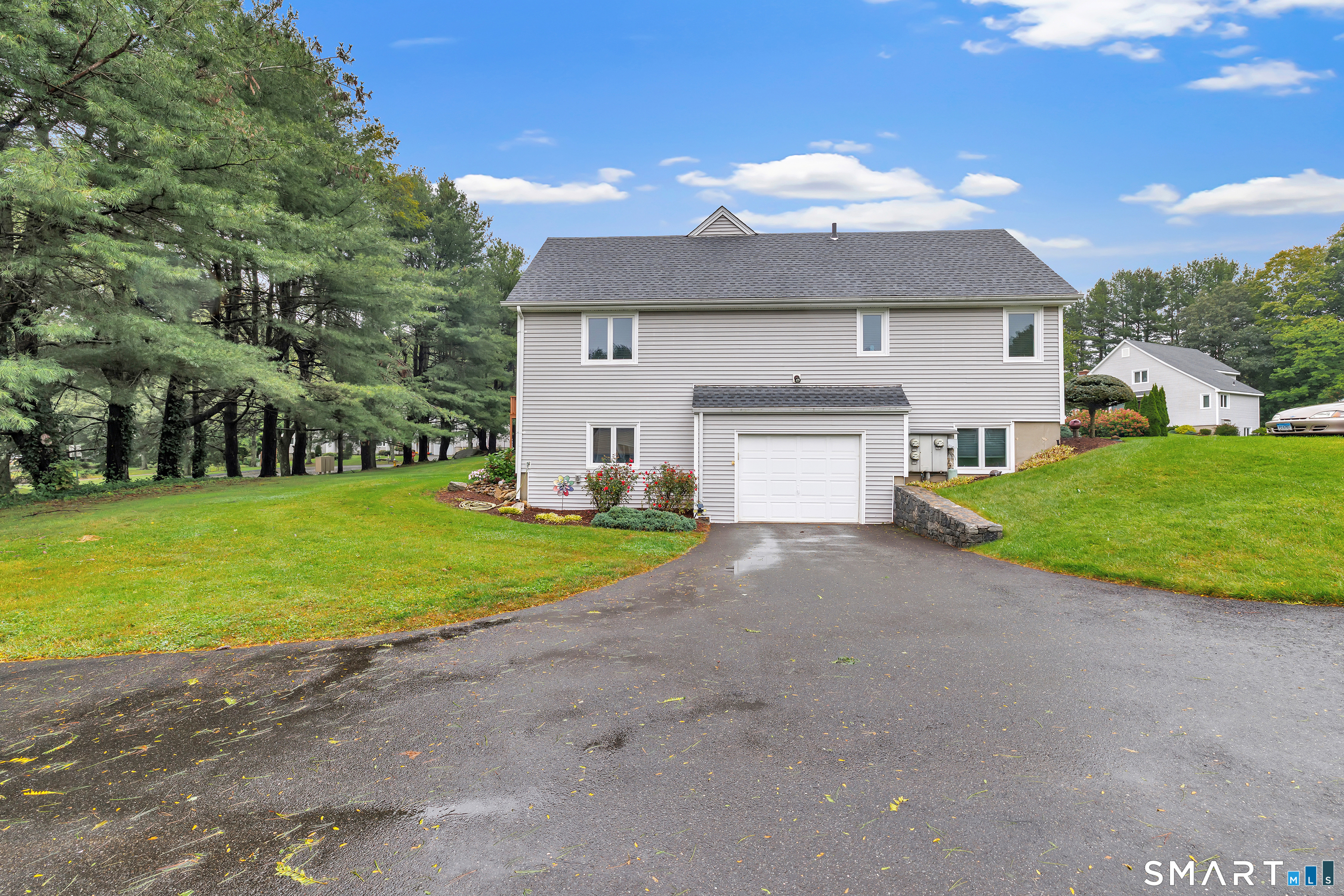 Two-story home with an attached garage, wet driveway, landscaped lawn, and mature trees.