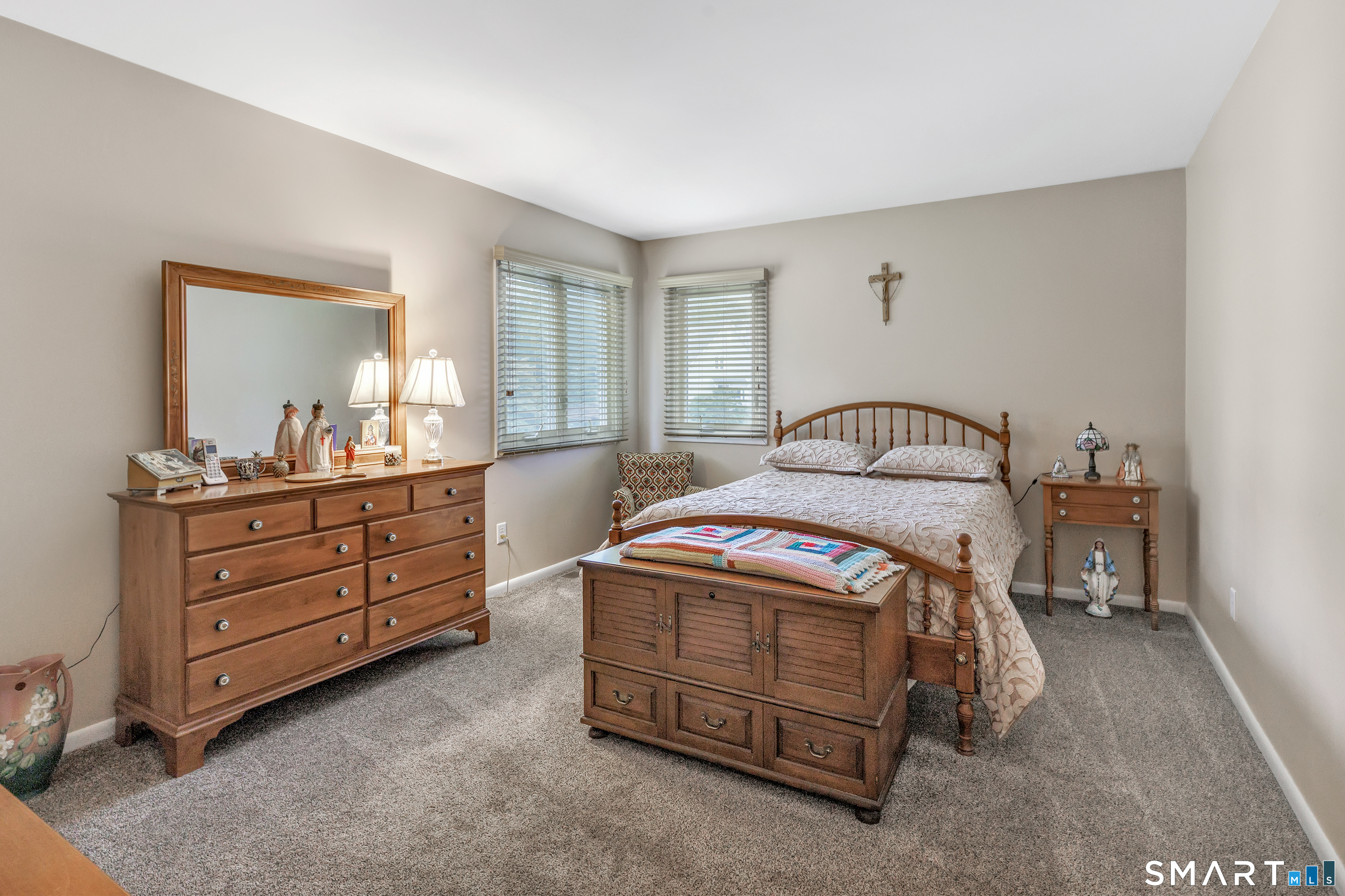 Furnished bedroom with a wooden bed, matching dresser, storage bench, and two windows with blinds.