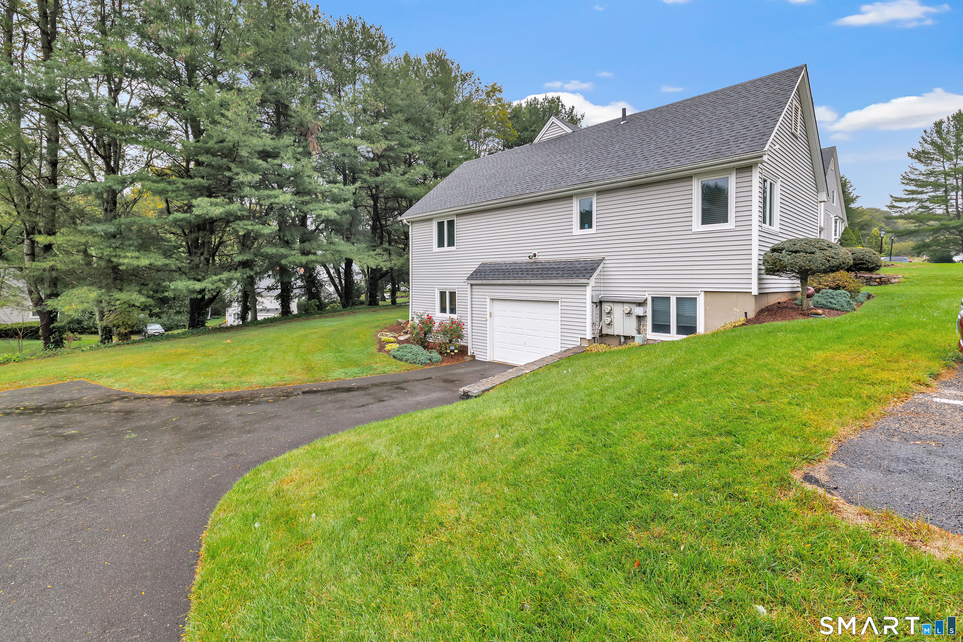Light-gray multi-level home with an attached garage, sloping lawn, curved driveway, and mature trees.
