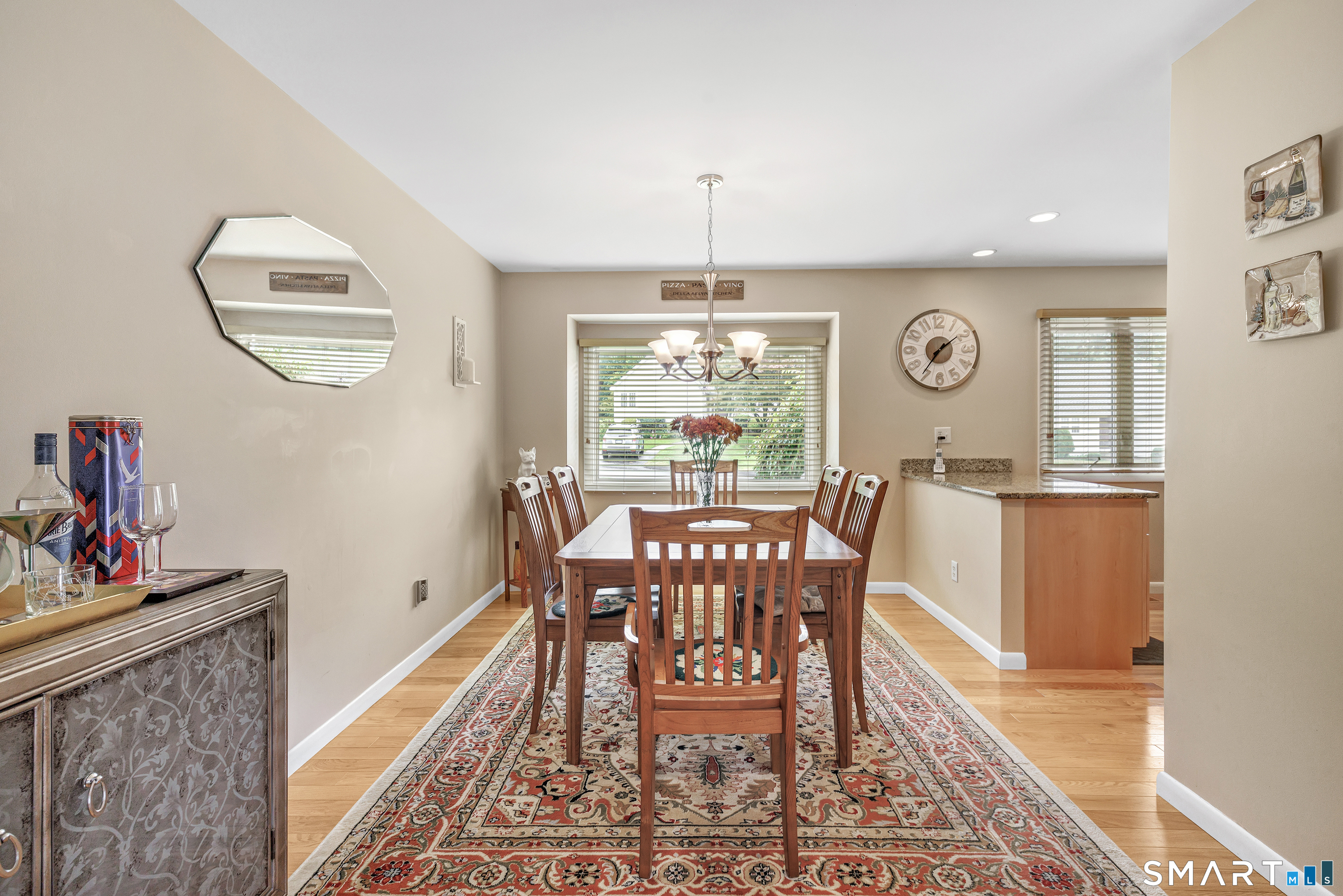 Furnished dining room with a wooden table, chandelier, patterned rug, and windows overlooking greenery.