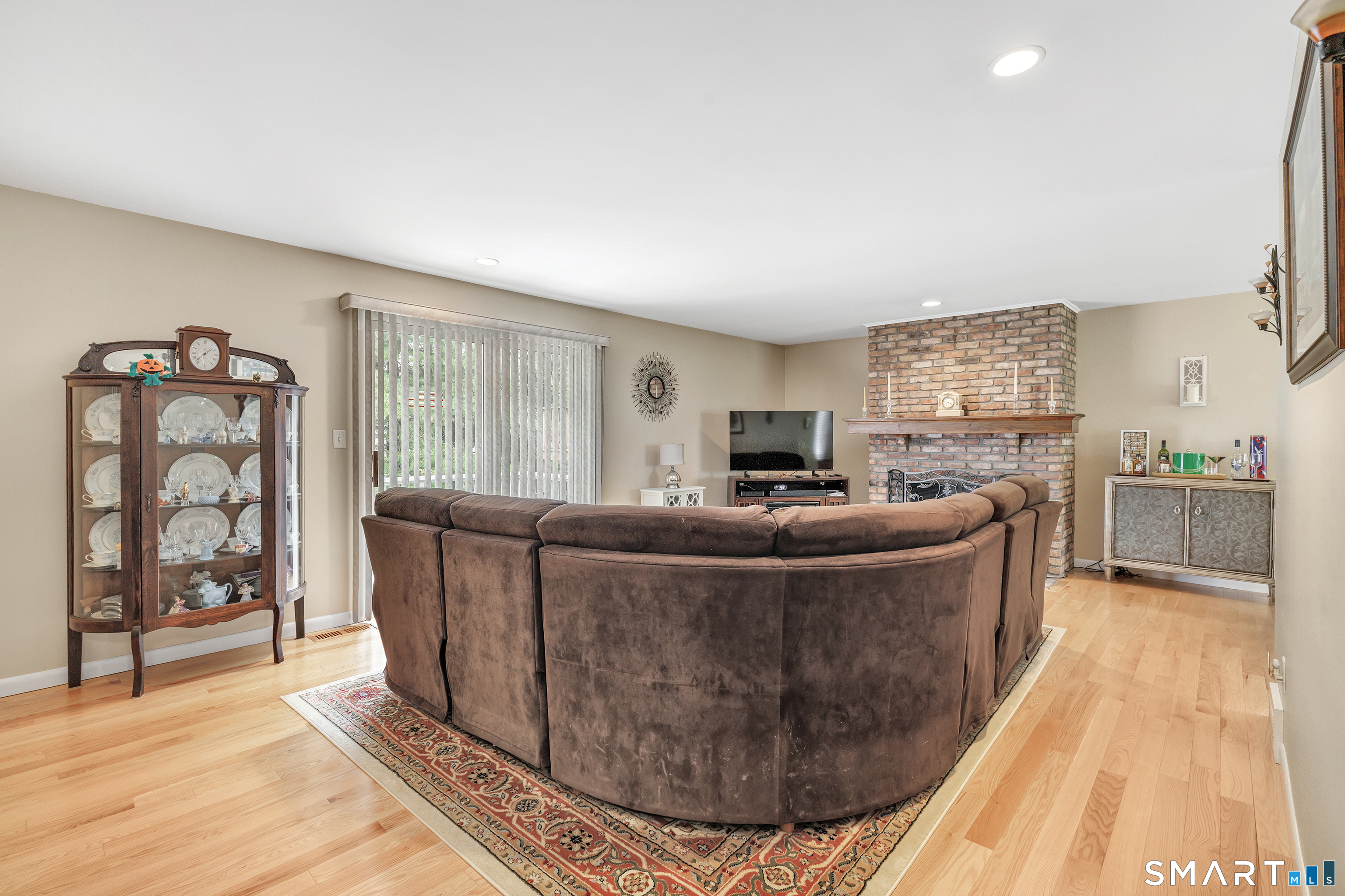 Living room with a curved brown sectional, brick fireplace, hardwood floors, and glass-front china cabinet.