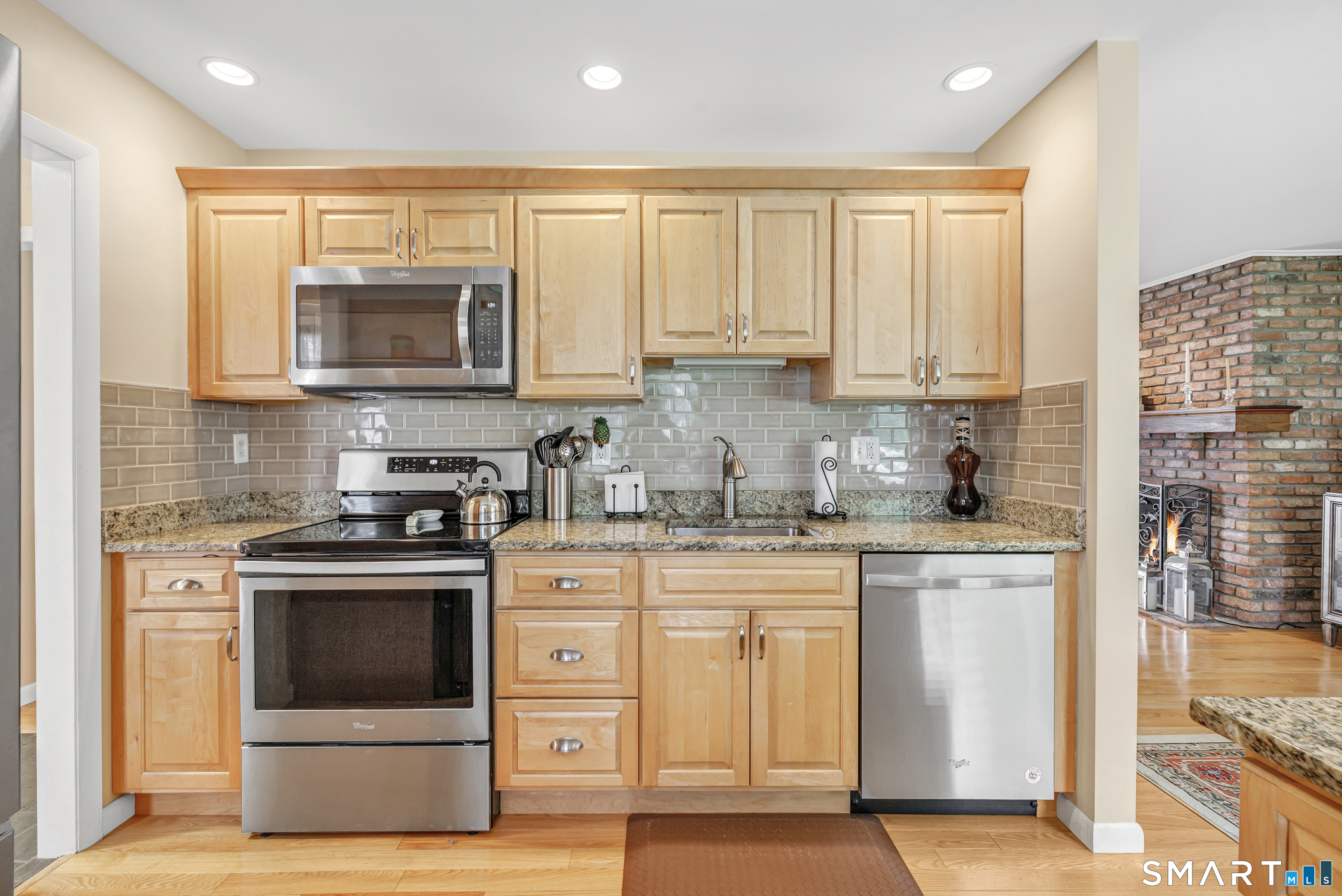 Kitchen with light wood cabinets, granite counters, stainless steel appliances, and a brick fireplace in the adjoining room.