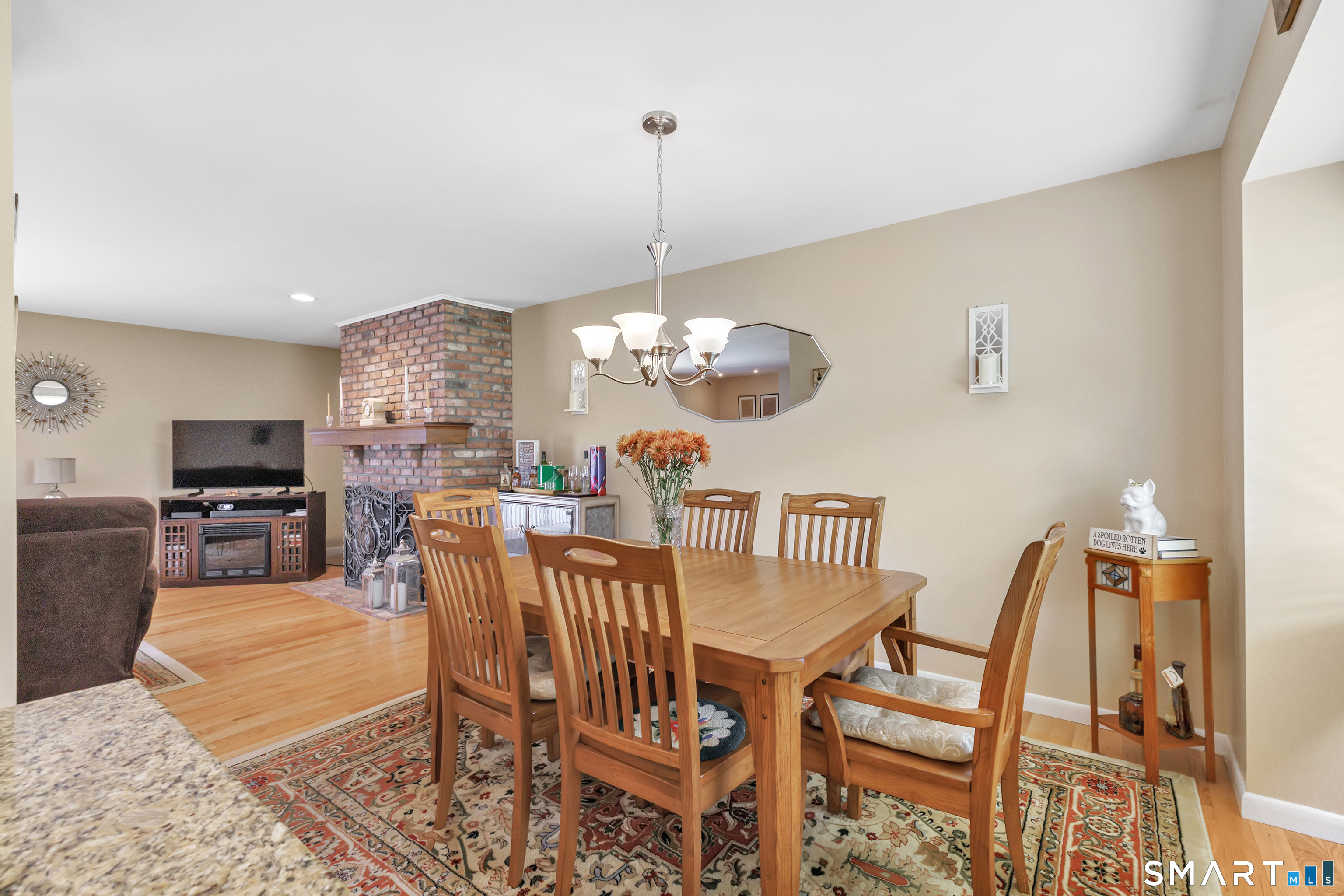 Dining area with a wooden table and chairs, patterned rug, and brick fireplace in the adjoining living room.