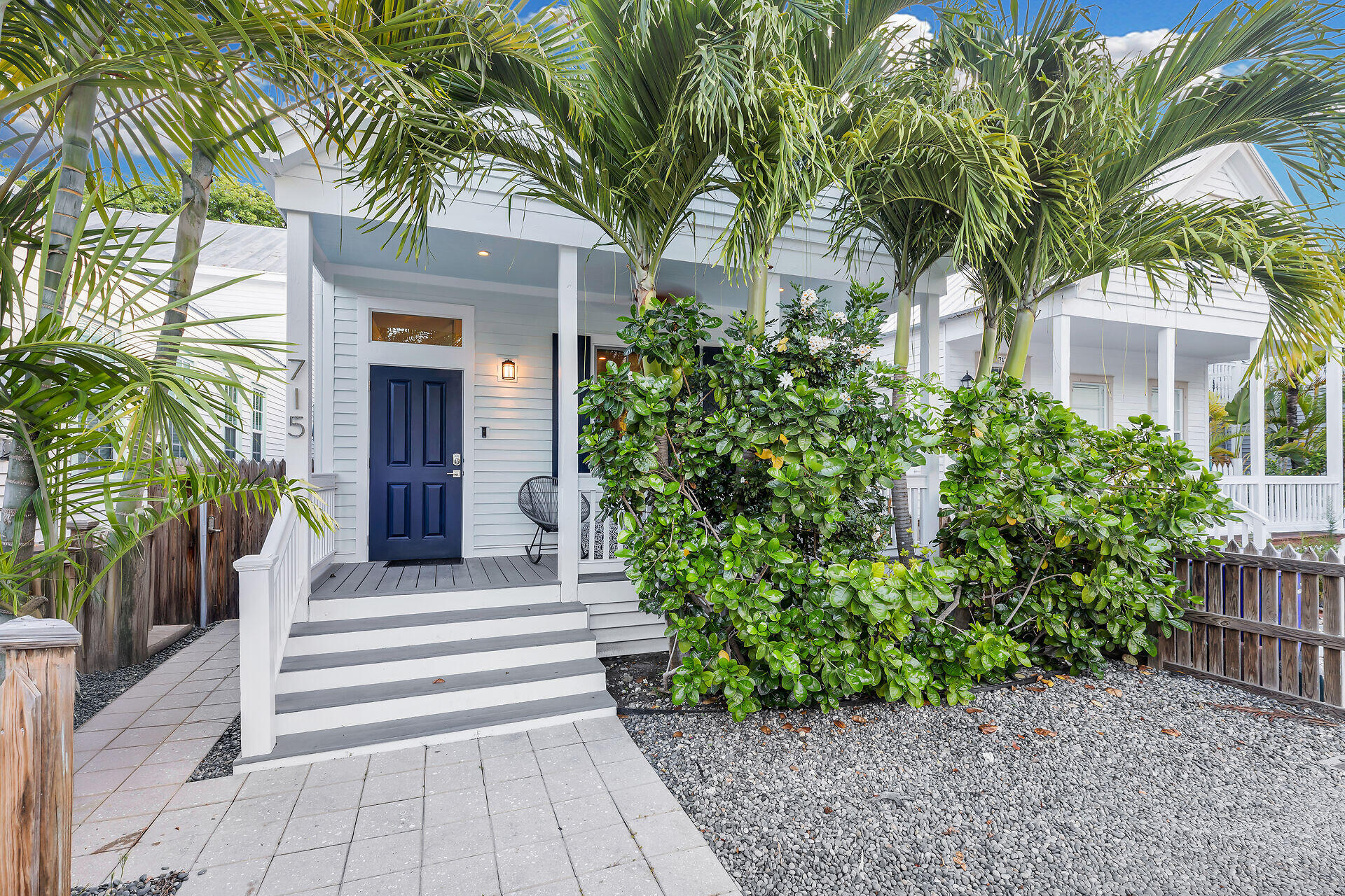 White home with a blue front door and porch framed by palms and lush shrubs.