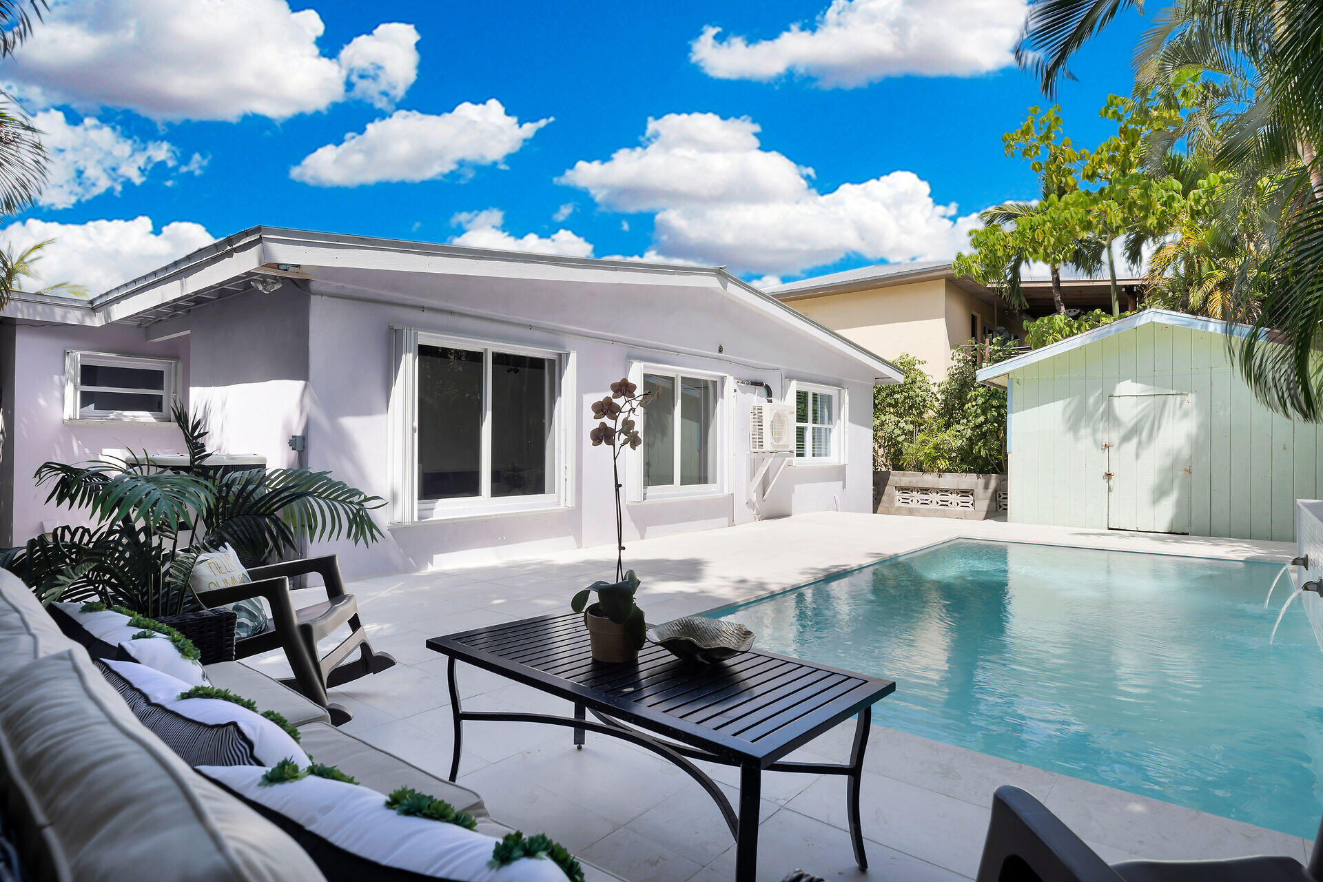 Backyard pool beside a single-story home, with lounge seating, tropical plants, and a pale green shed.