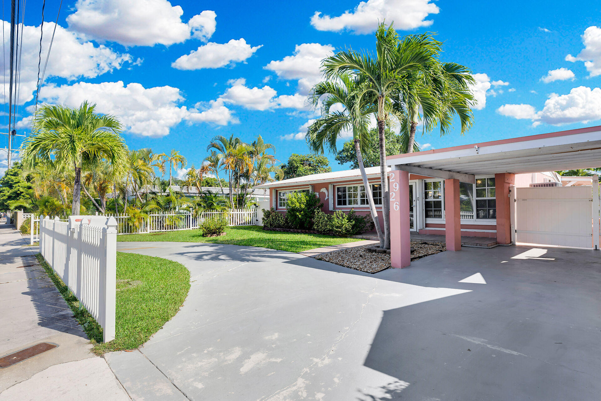 Single-story pink home with a covered carport, white picket fence, and palm trees.