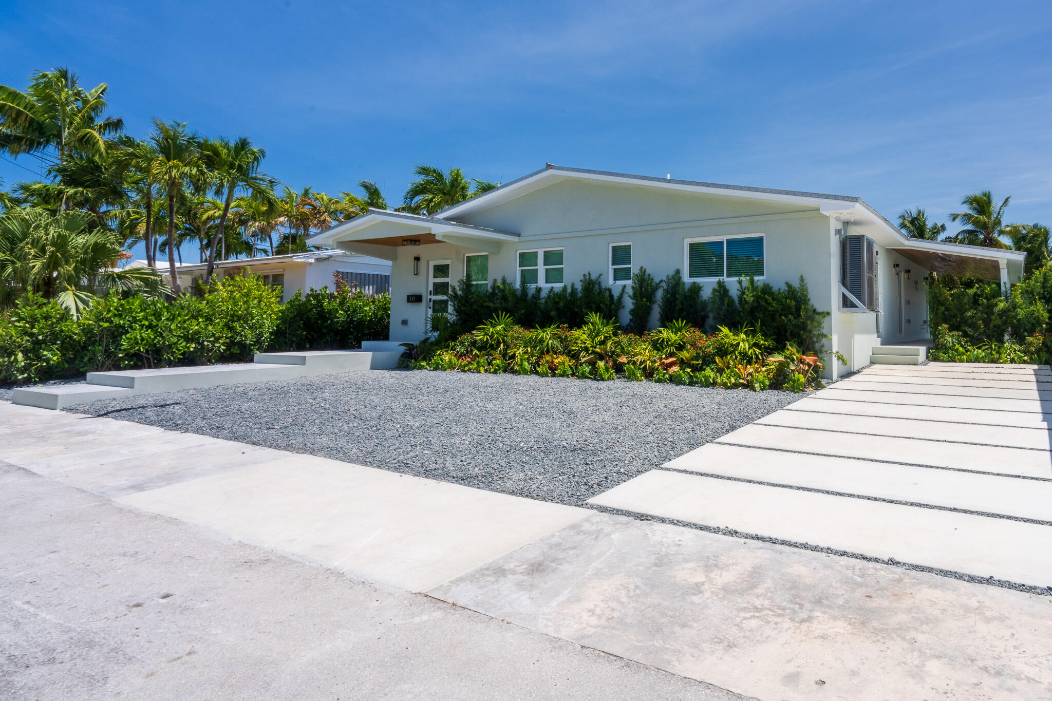 White single-story home with tropical landscaping, a gravel yard, and broad concrete stepping slabs.
