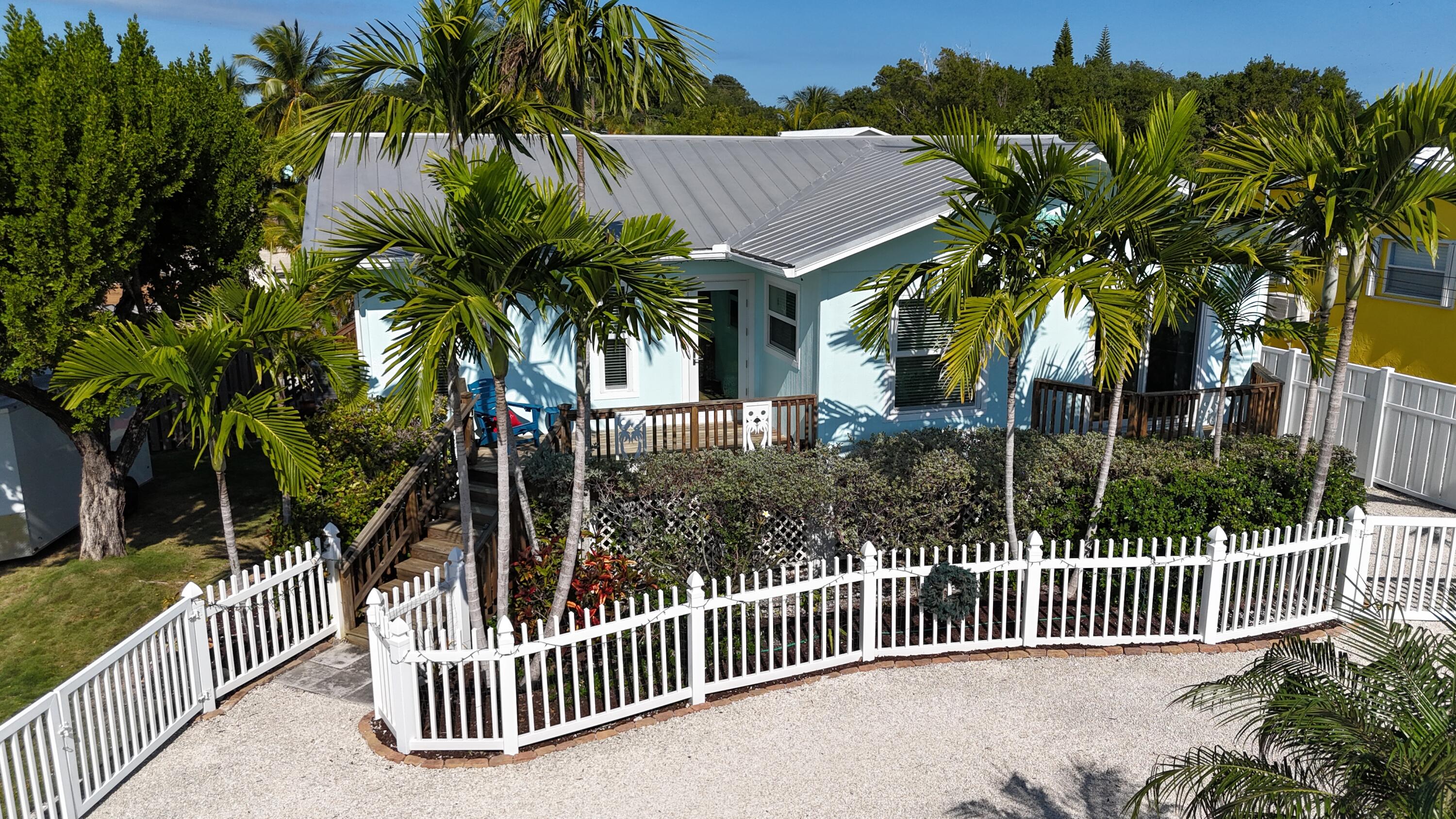Light-blue house with a metal roof, wooden porch, tropical palms, and white picket fence.