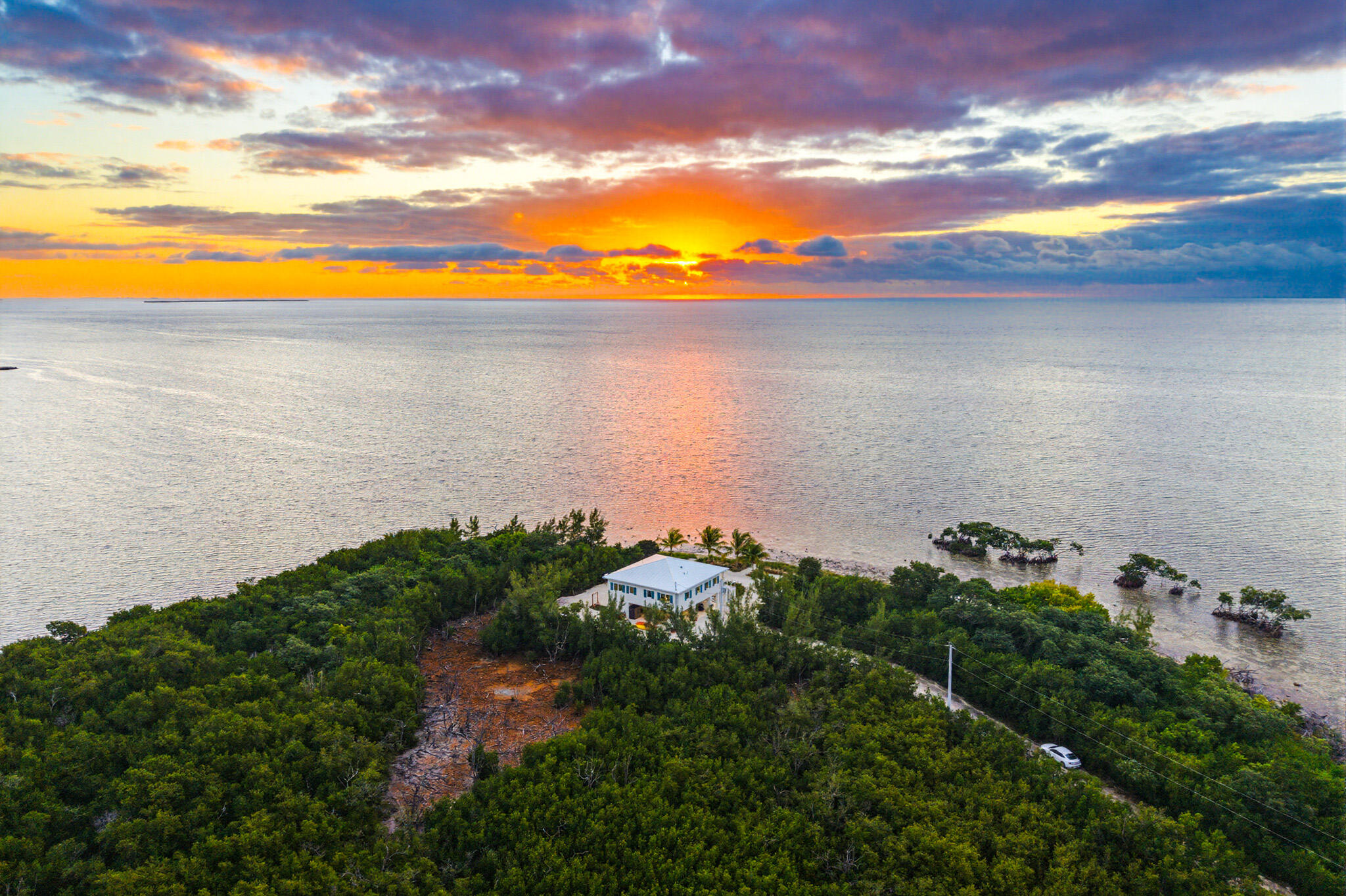 Aerial view of a white coastal home amid greenery, with mangrove islets and sunset over the water.