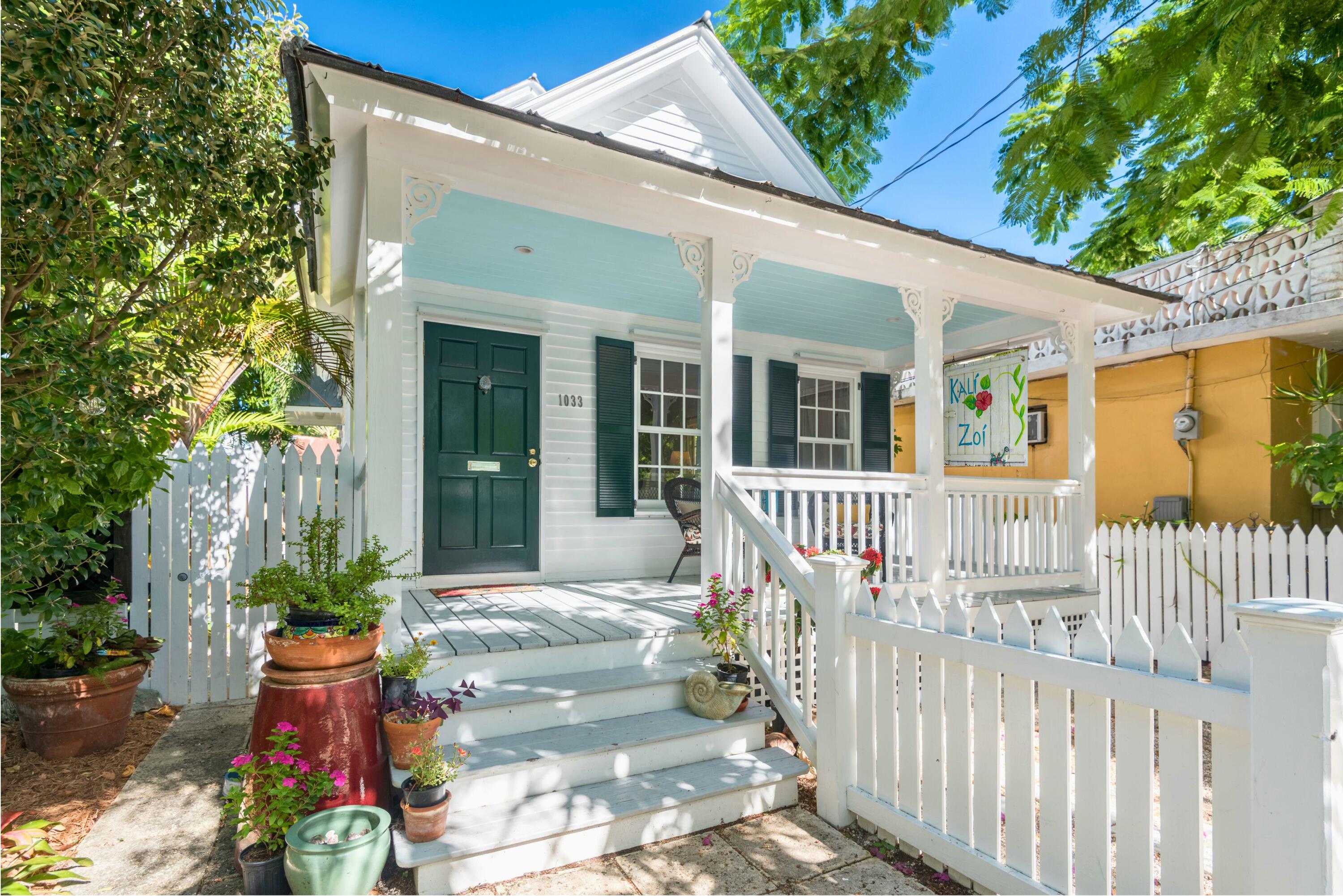 White house with a covered porch, pale blue ceiling, dark green door, and flower-filled tropical landscaping.