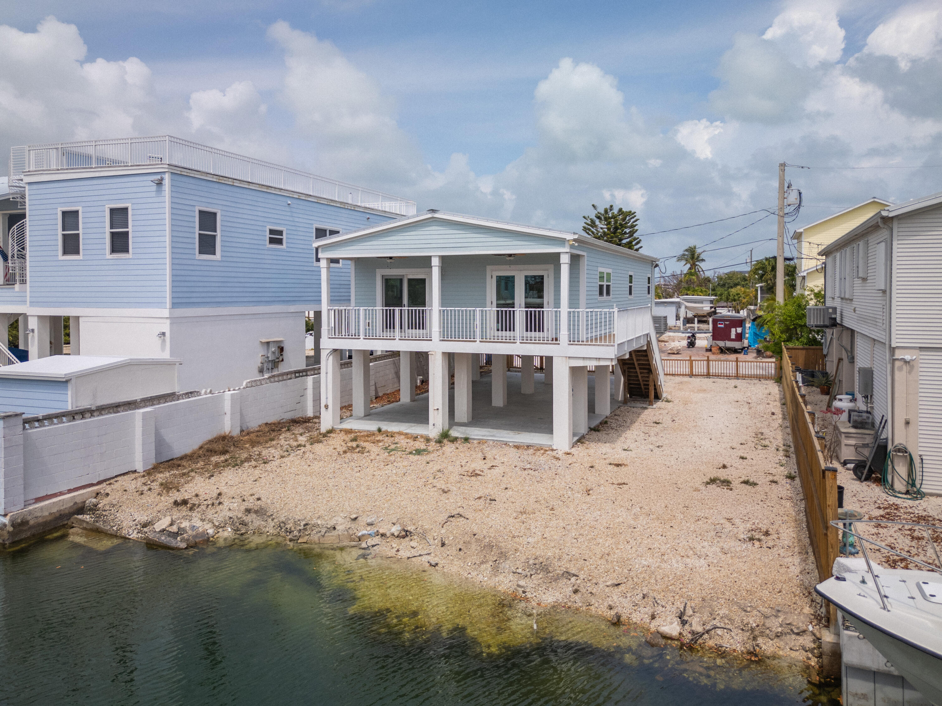 Elevated light-blue waterfront home with a covered deck and gravel shoreline.