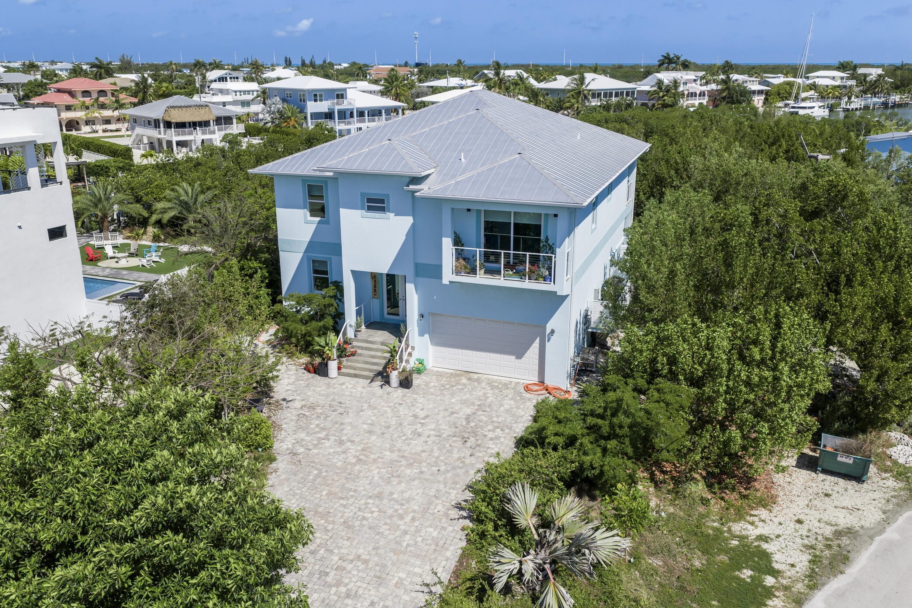 Blue-and-white two-story home with a metal roof, broad paved driveway, and tropical landscaping.
