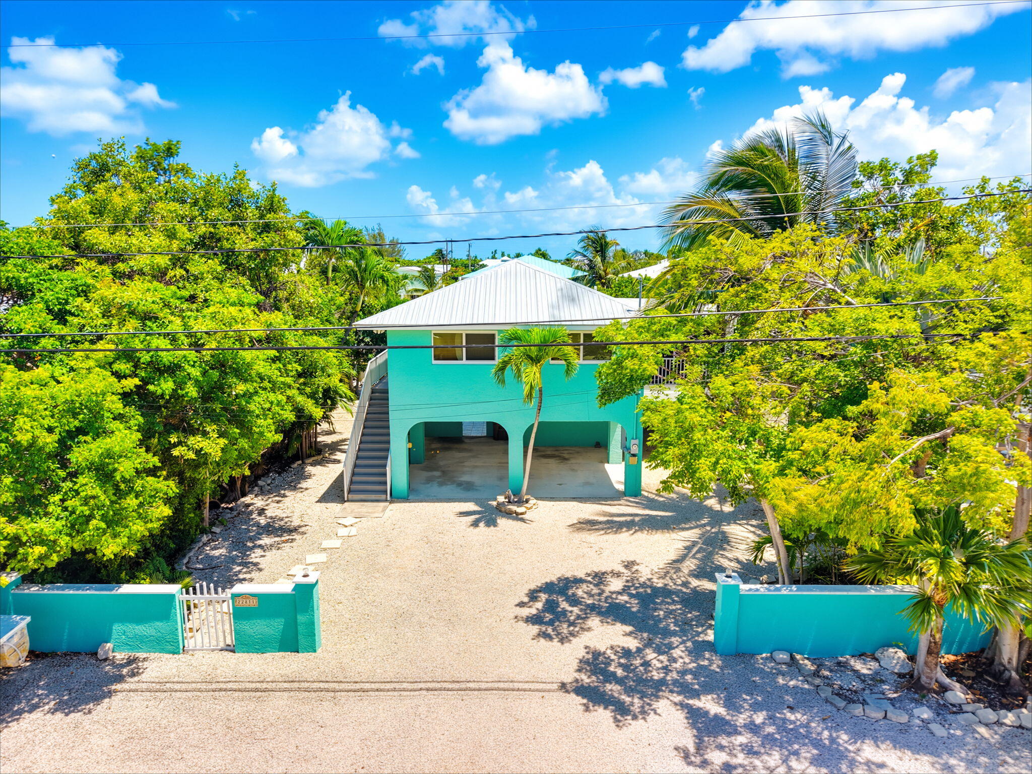 Turquoise raised house with a white roof, open ground-level parking, and a gravel driveway framed by tropical trees.