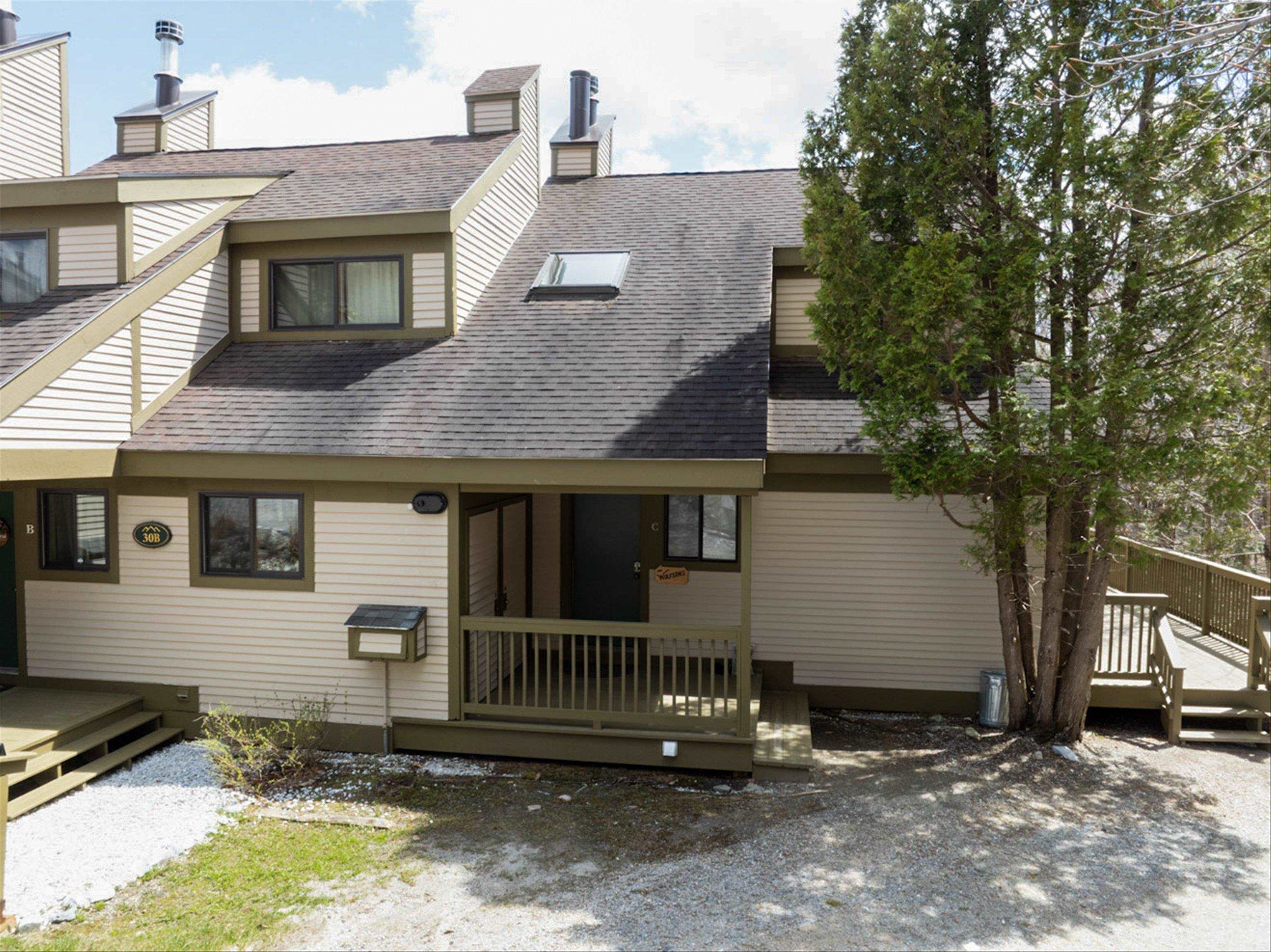 Cream condominium exterior with a covered porch, dark shingle roof, and wooden deck beside a large tree.