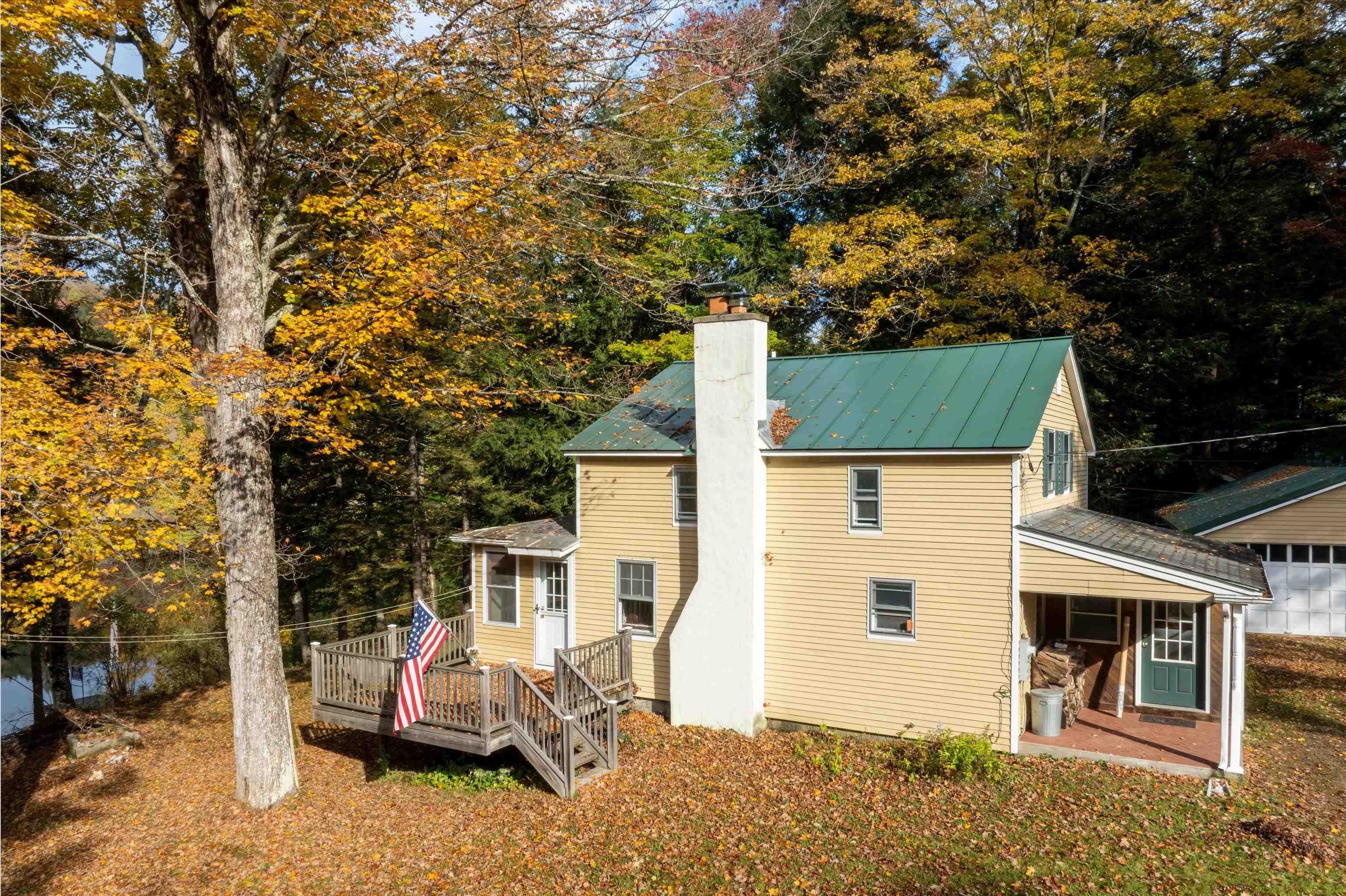 Yellow two-story home with a green metal roof, white chimney, and deck among autumn trees.