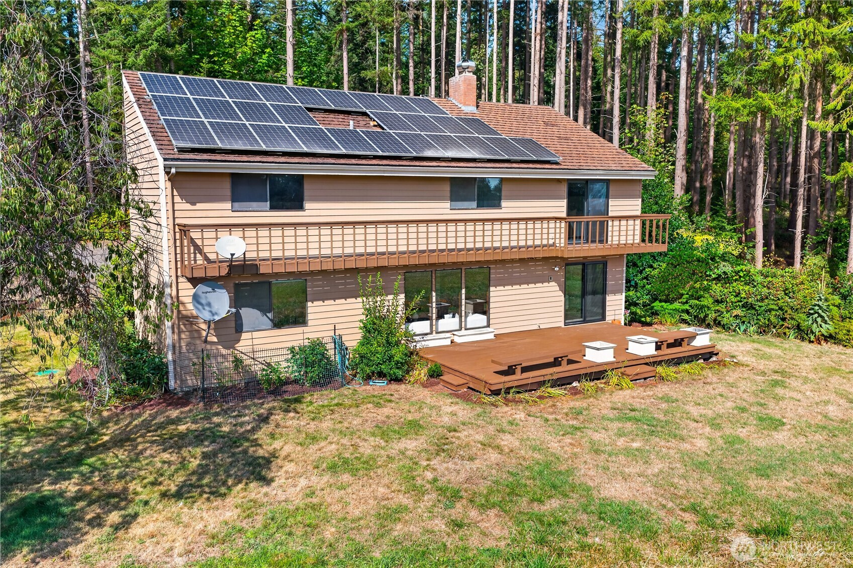 House rear with rooftop solar panels, a long balcony, and a broad deck among tall trees.