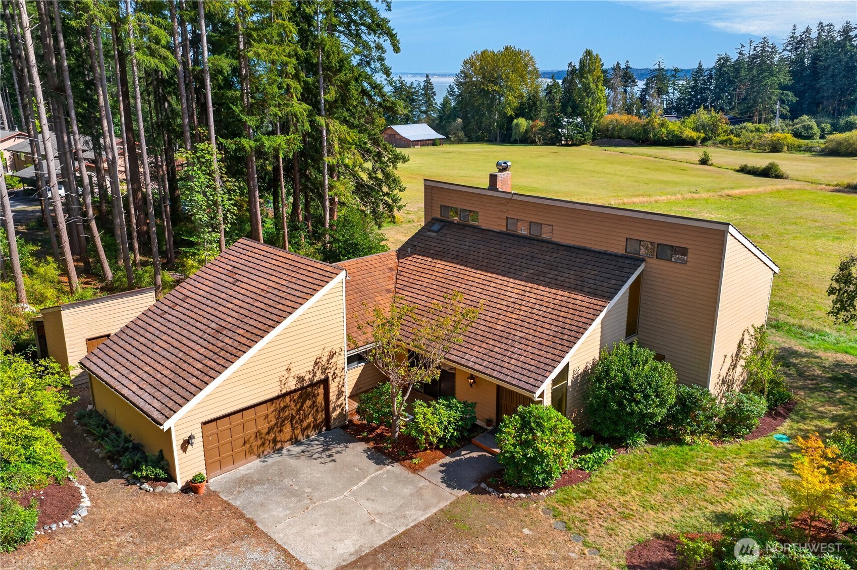 Aerial view of a tan home with an attached garage, broad lawn, surrounding trees, and distant water.