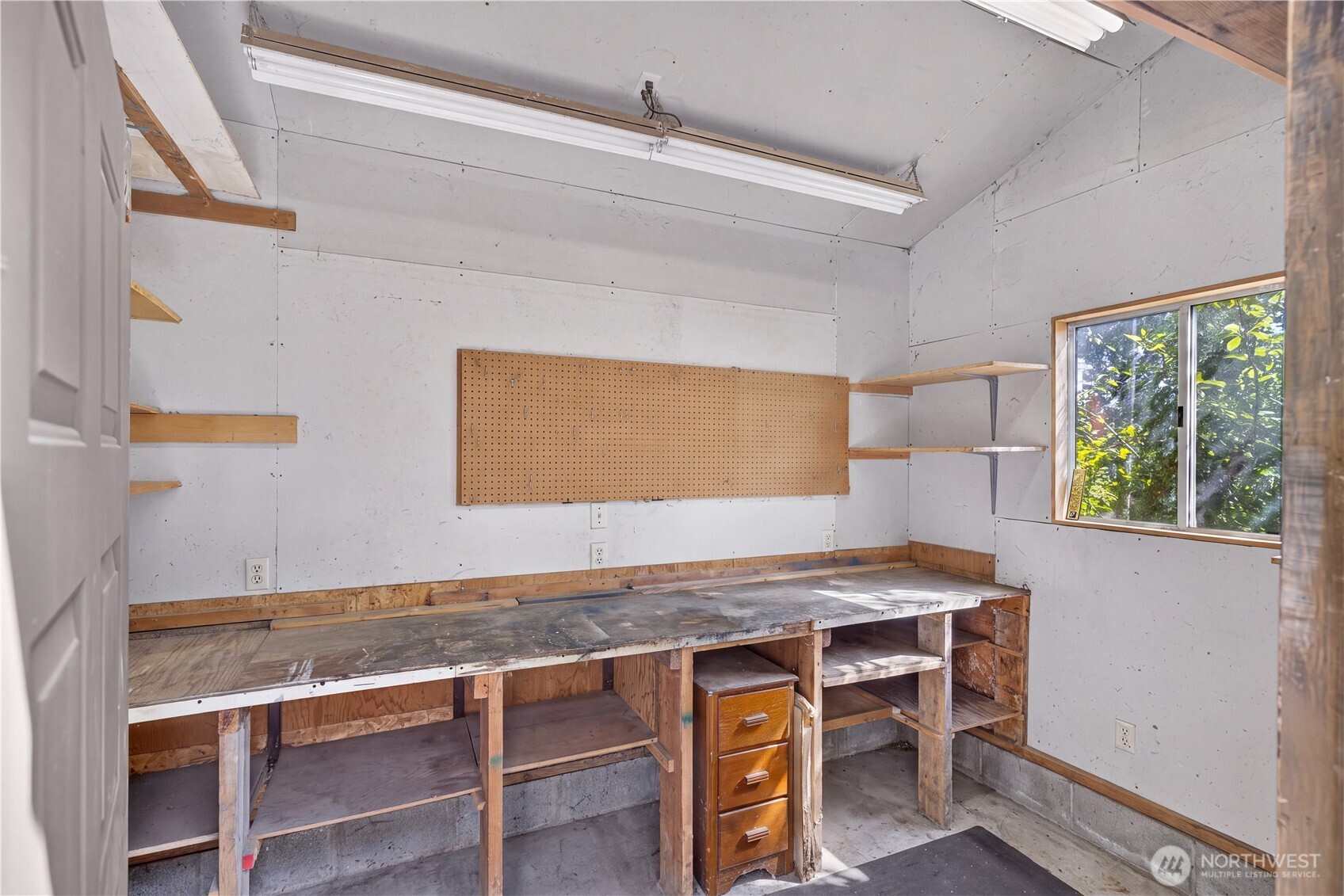 Workshop with a long built-in workbench, pegboard panel, open shelves, and a window overlooking trees.