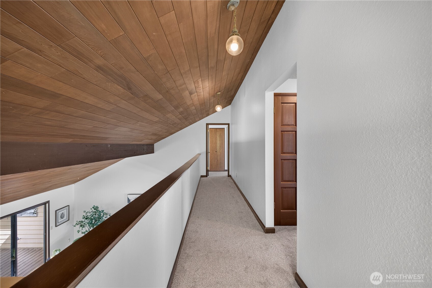 Upstairs hallway with a sloped wood-plank ceiling, carpeted floor, and railing overlooking the lower level.