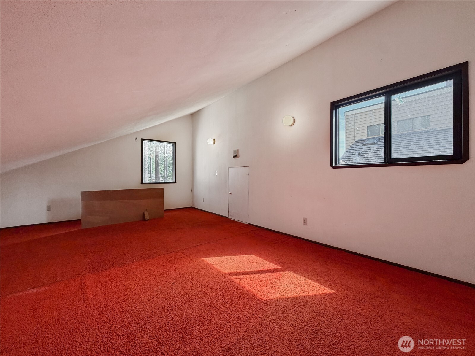 Empty carpeted loft with a sloped ceiling, two windows, and sunlight on the red floor.