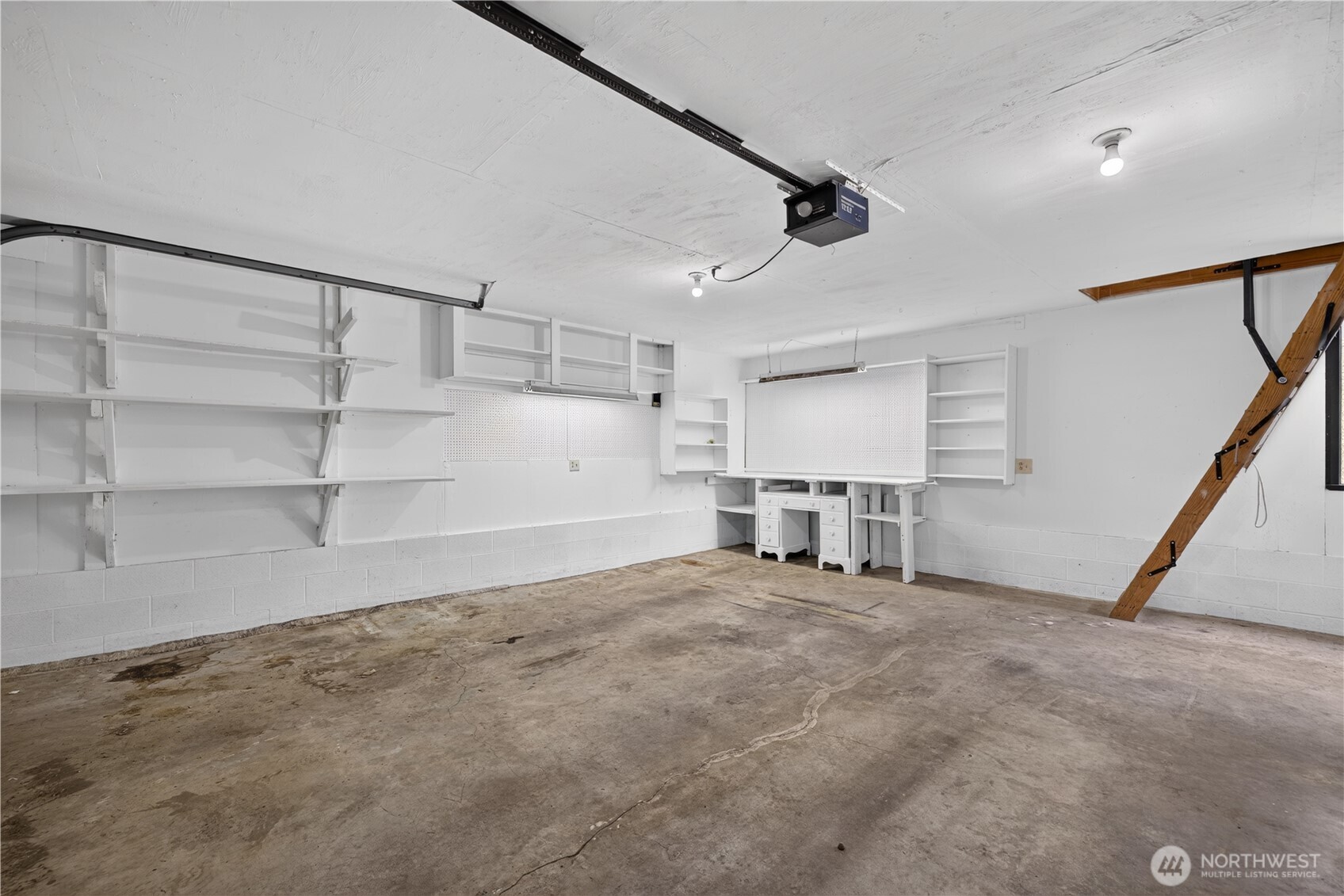 Empty garage with extensive white shelving, a built-in workbench, and a folded wooden attic ladder.