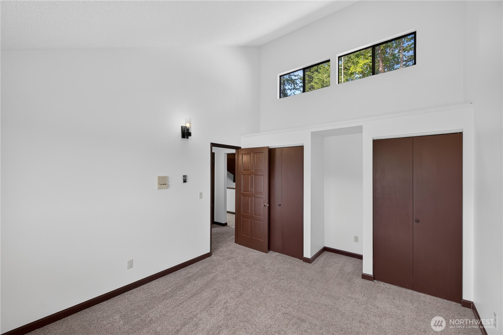Empty bedroom with gray carpet, dark wood closet doors, and high windows overlooking trees.