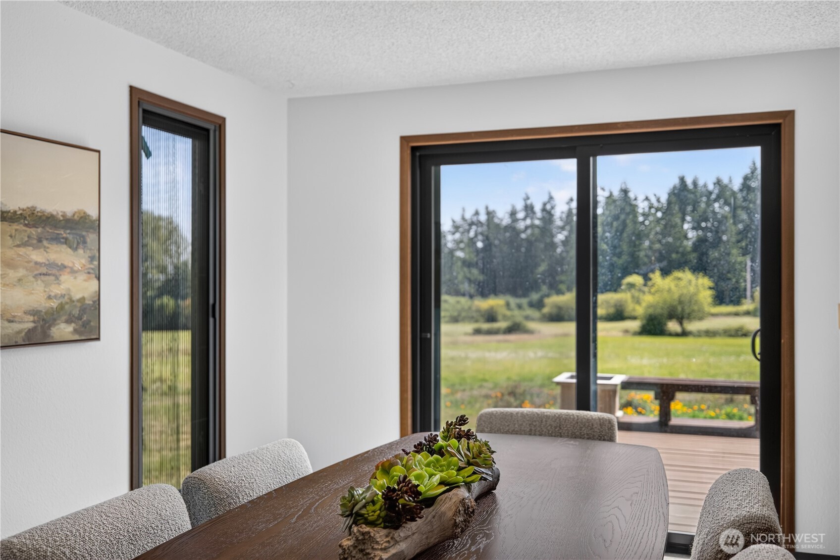 Dining room with a long wood table overlooking a deck and tree-lined meadow through sliding glass doors.
