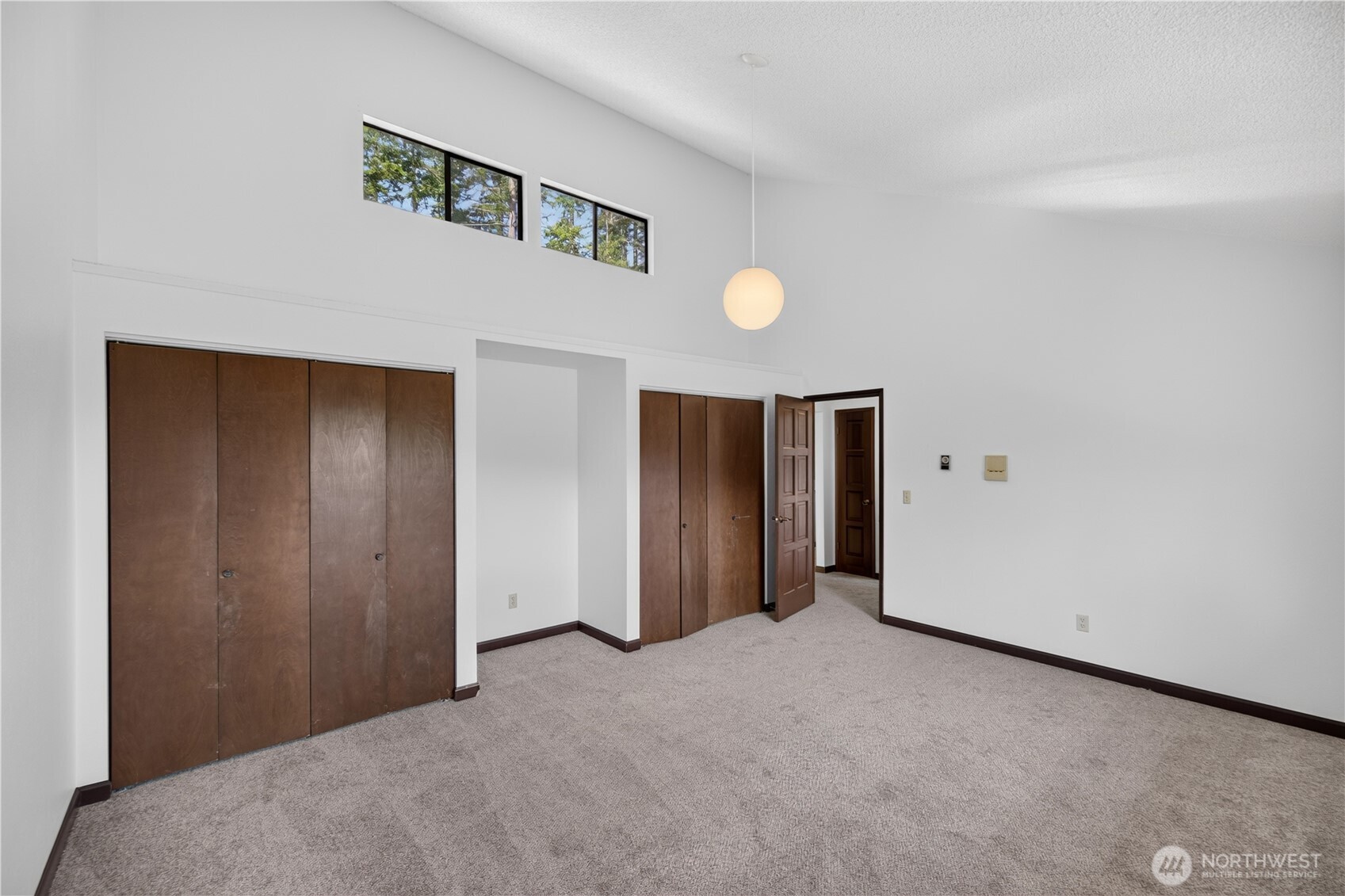 Empty bedroom with carpet, brown closet doors, high windows, and a round pendant light.