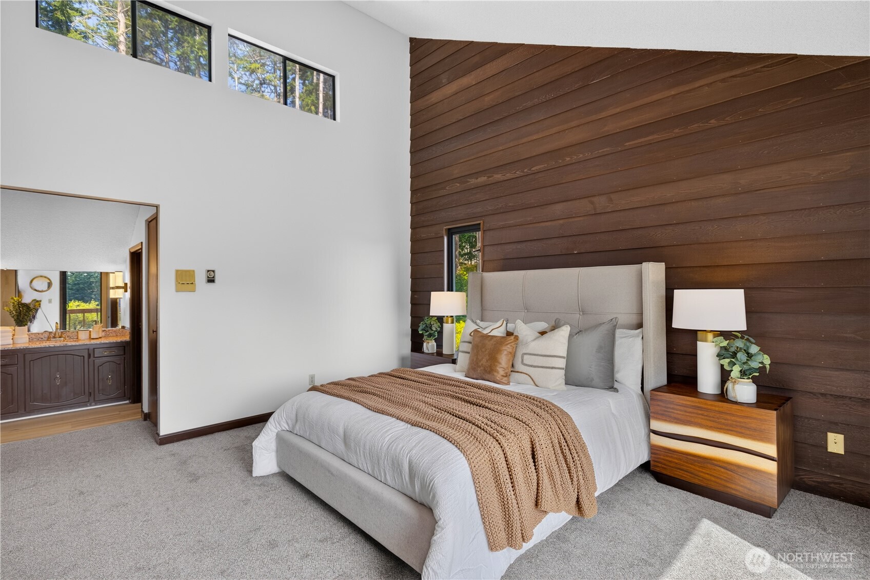 Staged bedroom with a tall wood accent wall, high windows, and an adjoining bathroom vanity.