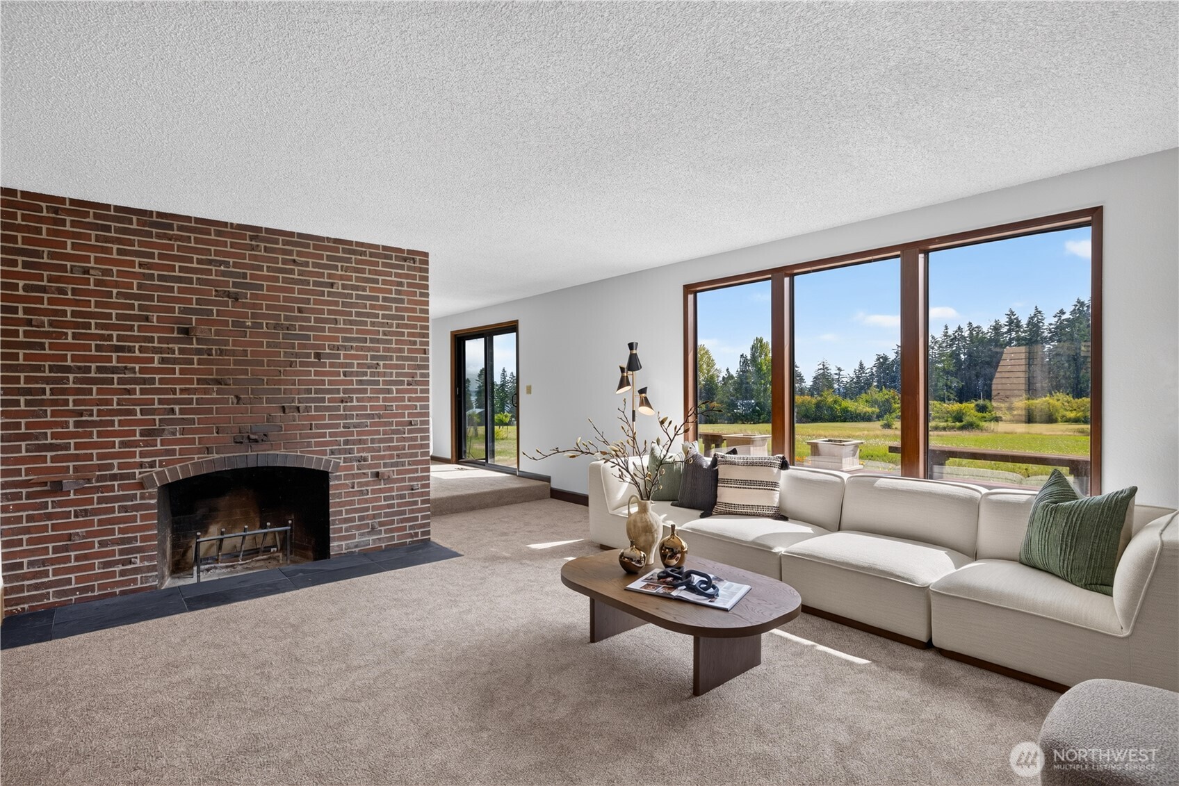 Living room with a brick fireplace, cream sectional, and large windows overlooking greenery.
