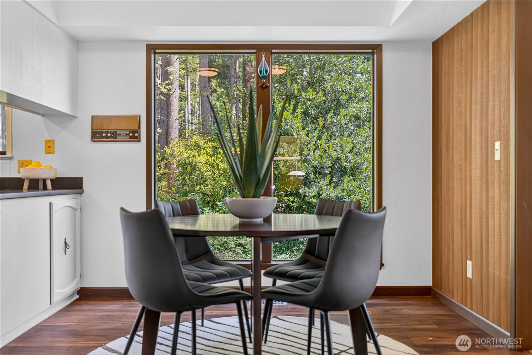 Dining area with gray chairs, a large potted plant, and windows overlooking trees.