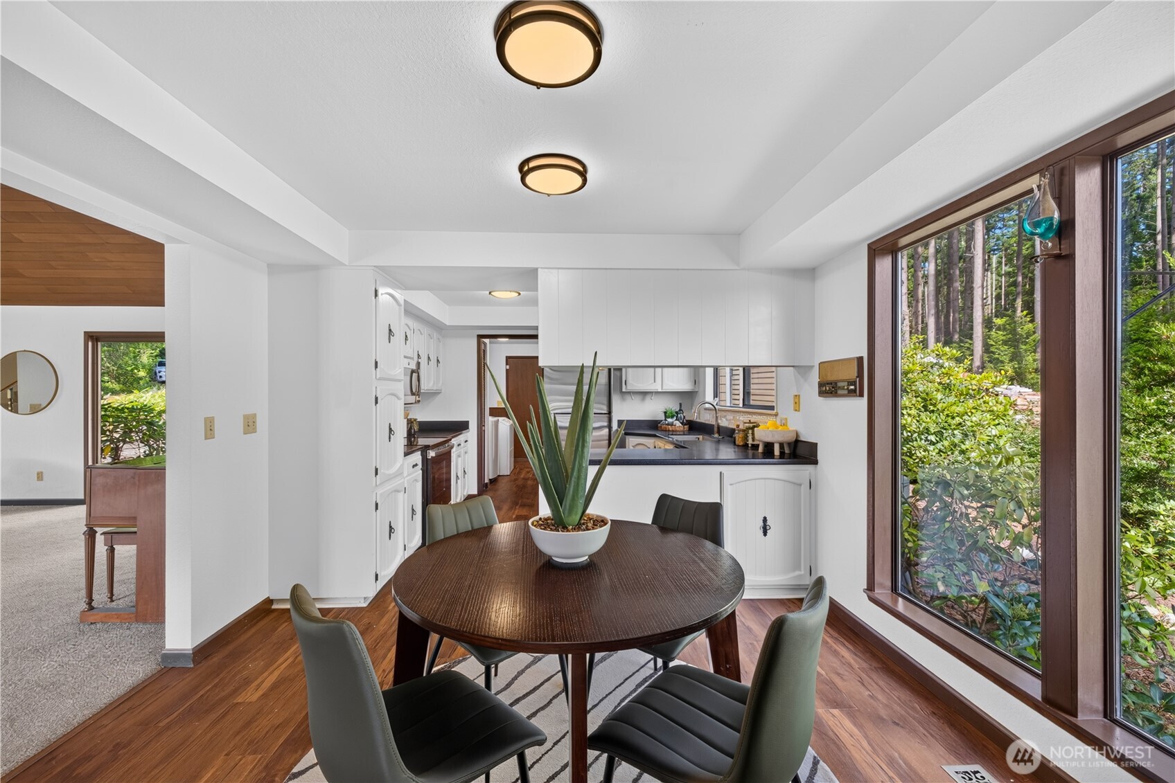 Dining area beside a white kitchen, with a round wood table and large garden-facing windows.
