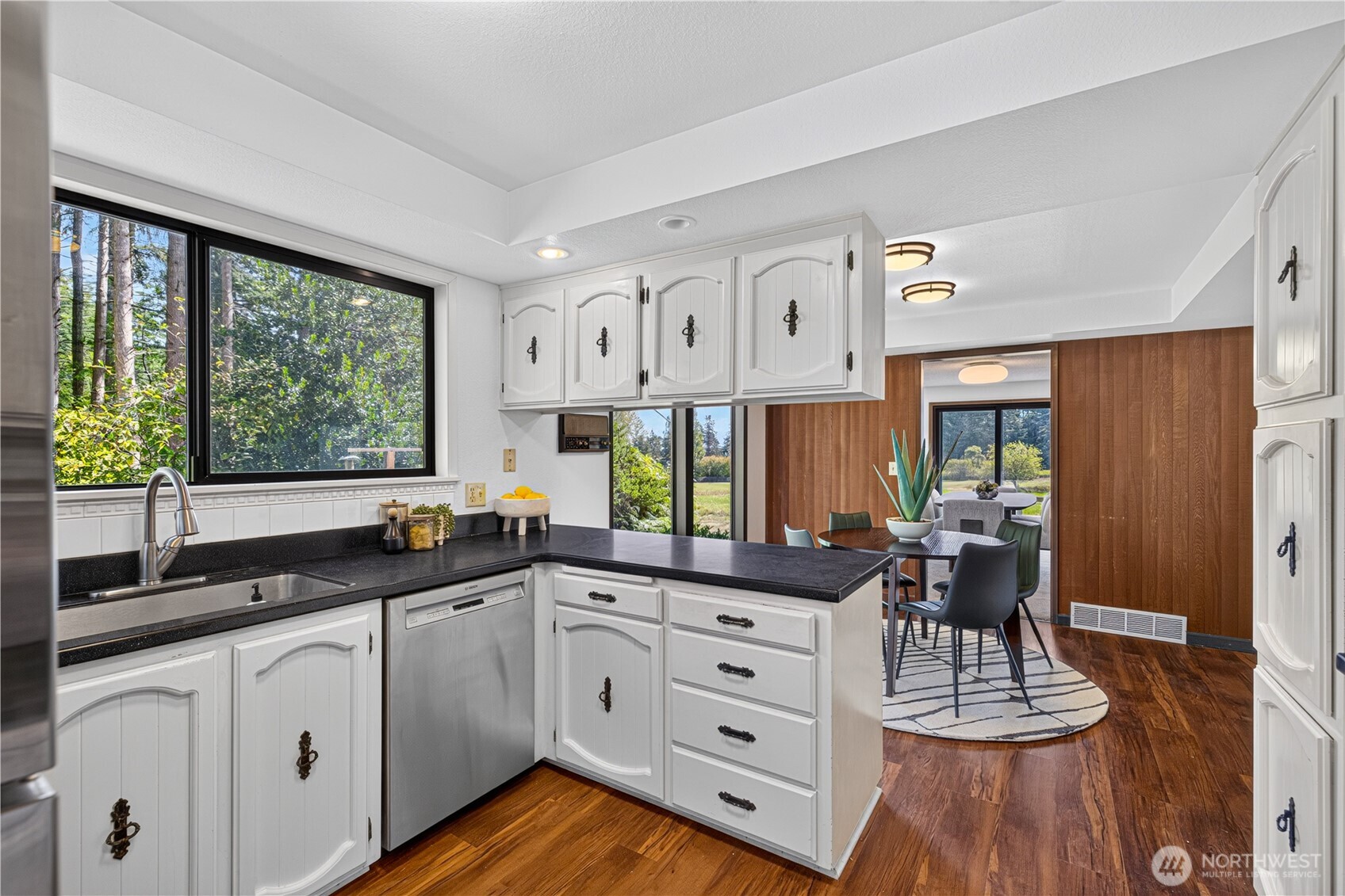 Kitchen with white cabinets and dark counters opening to a wood-paneled dining area.