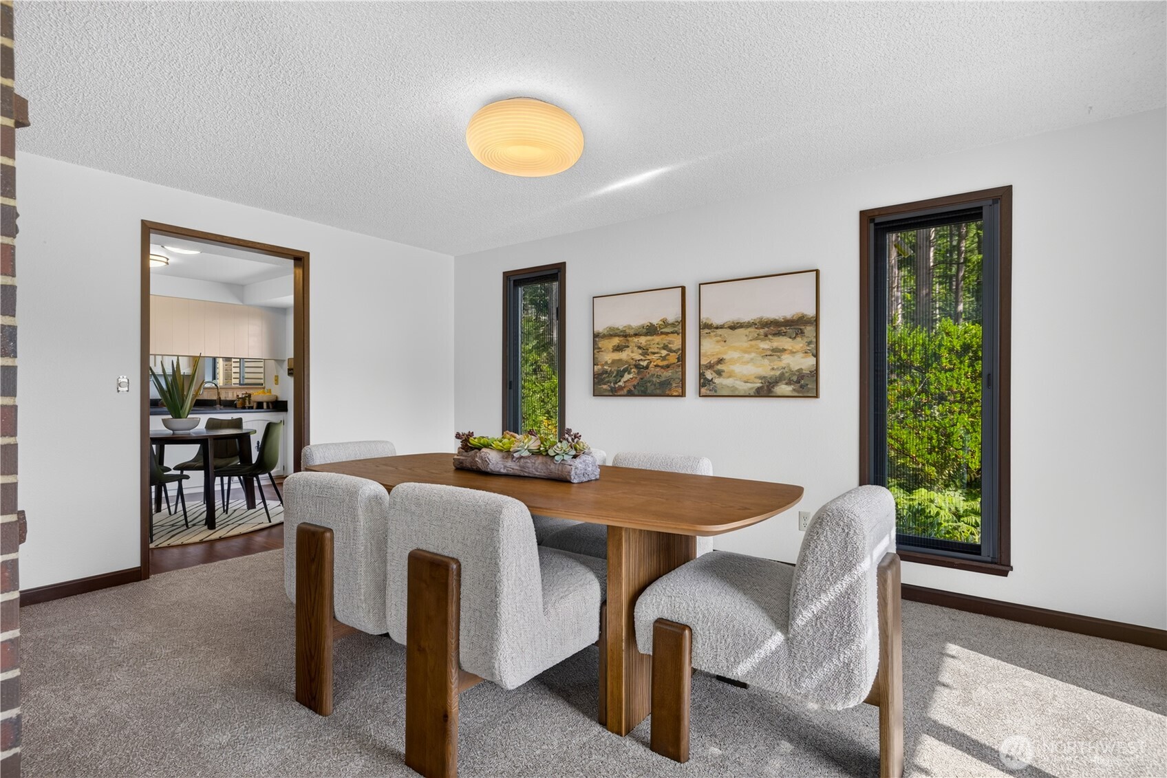 Dining room with a wooden table, upholstered chairs, and tall windows overlooking greenery.