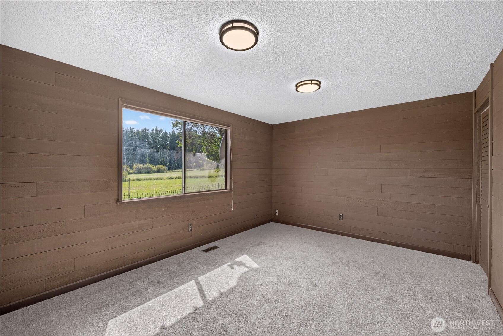 Empty bedroom with brown wood-paneled walls, gray carpet, and a sunny window overlooking a grassy yard.
