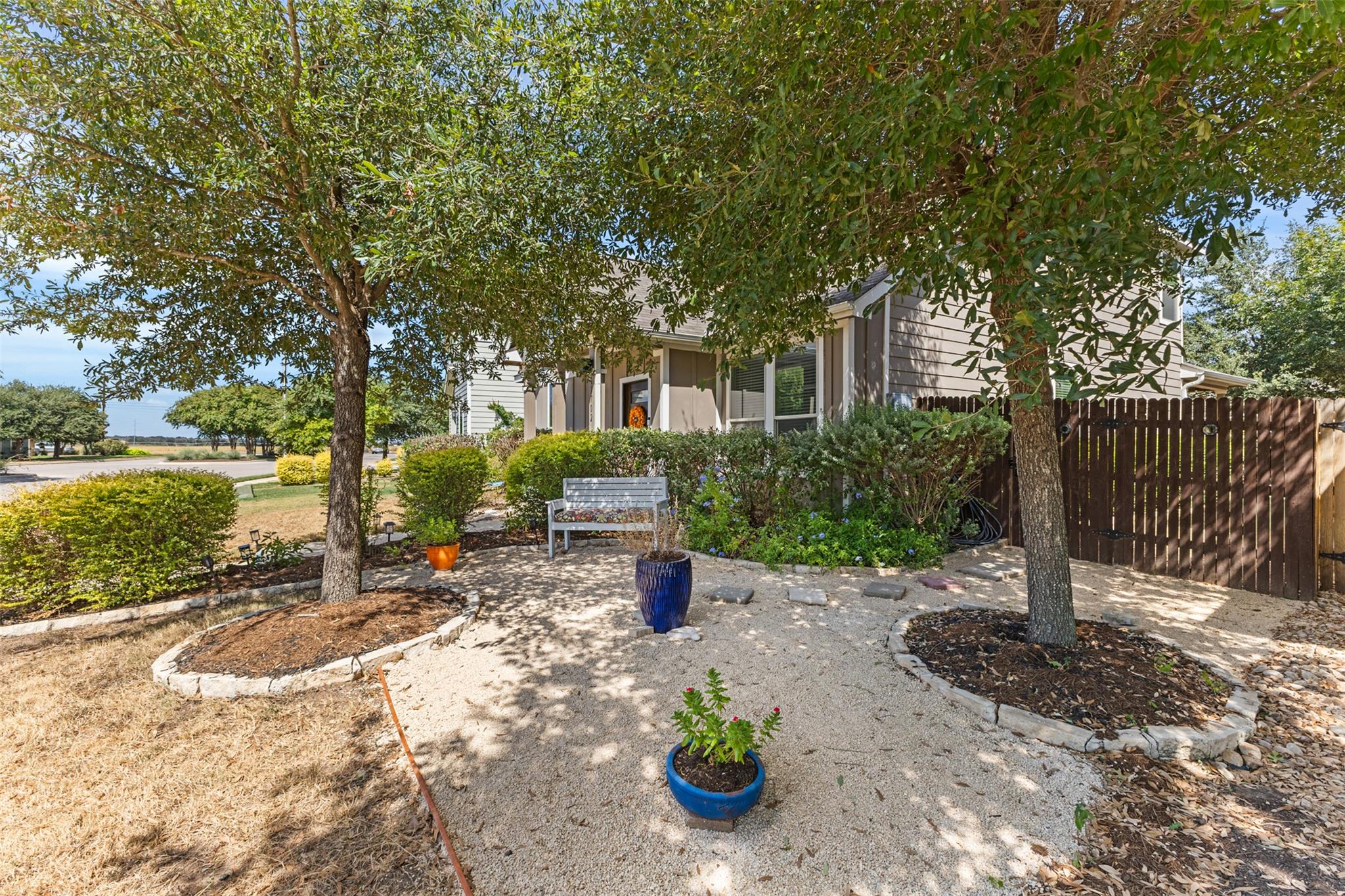 Home beside a shaded gravel patio with mature trees, a white bench, and potted plants.