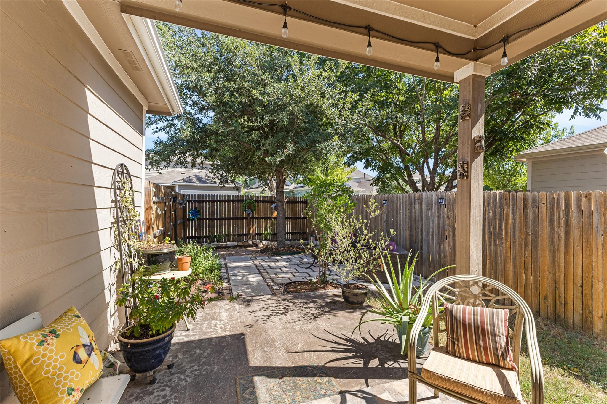 Covered backyard patio with string lights, potted plants, and a stone path leading to mature trees.