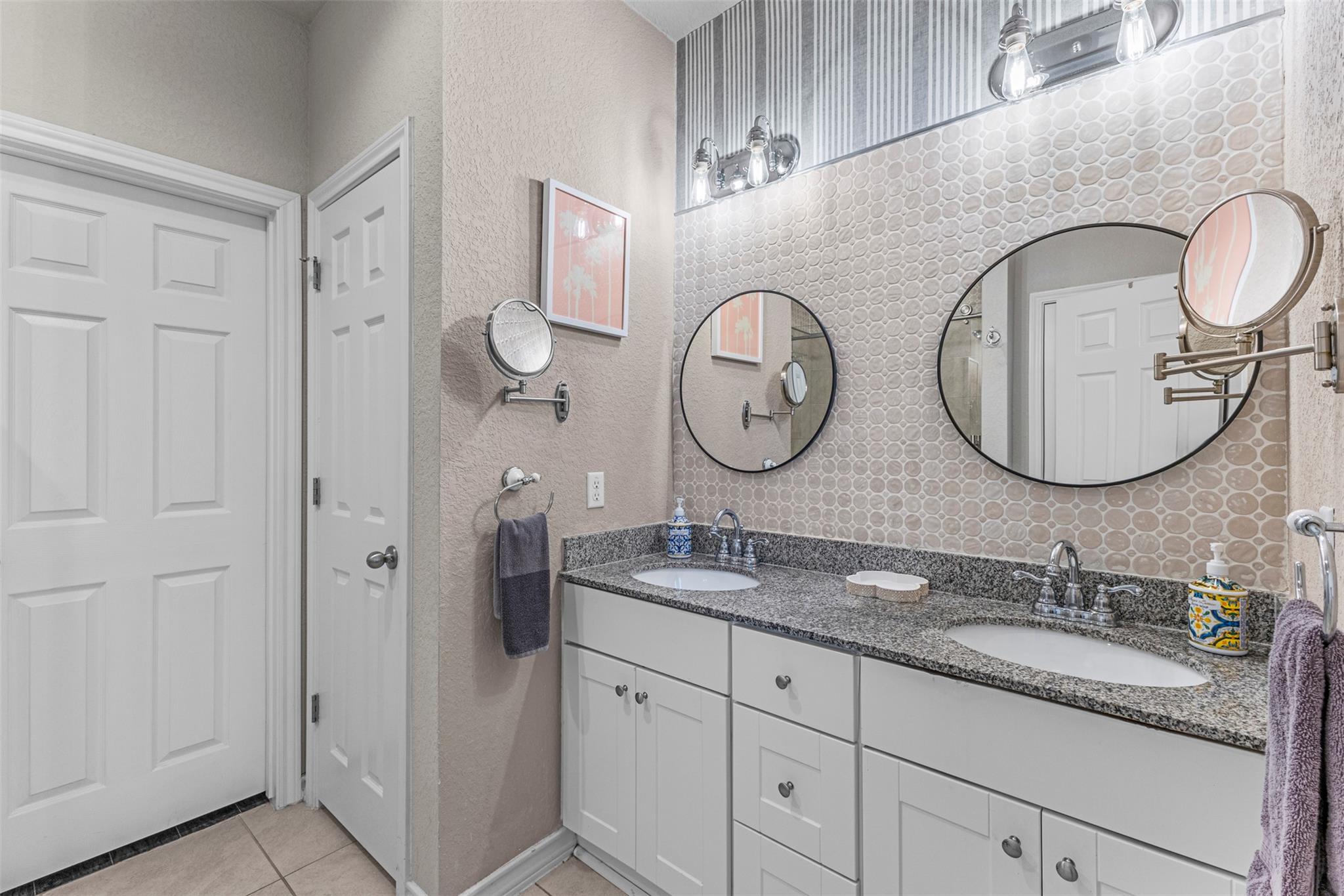 Bathroom with a double-sink vanity, granite countertop, round mirrors, and circular-pattern tile backsplash.