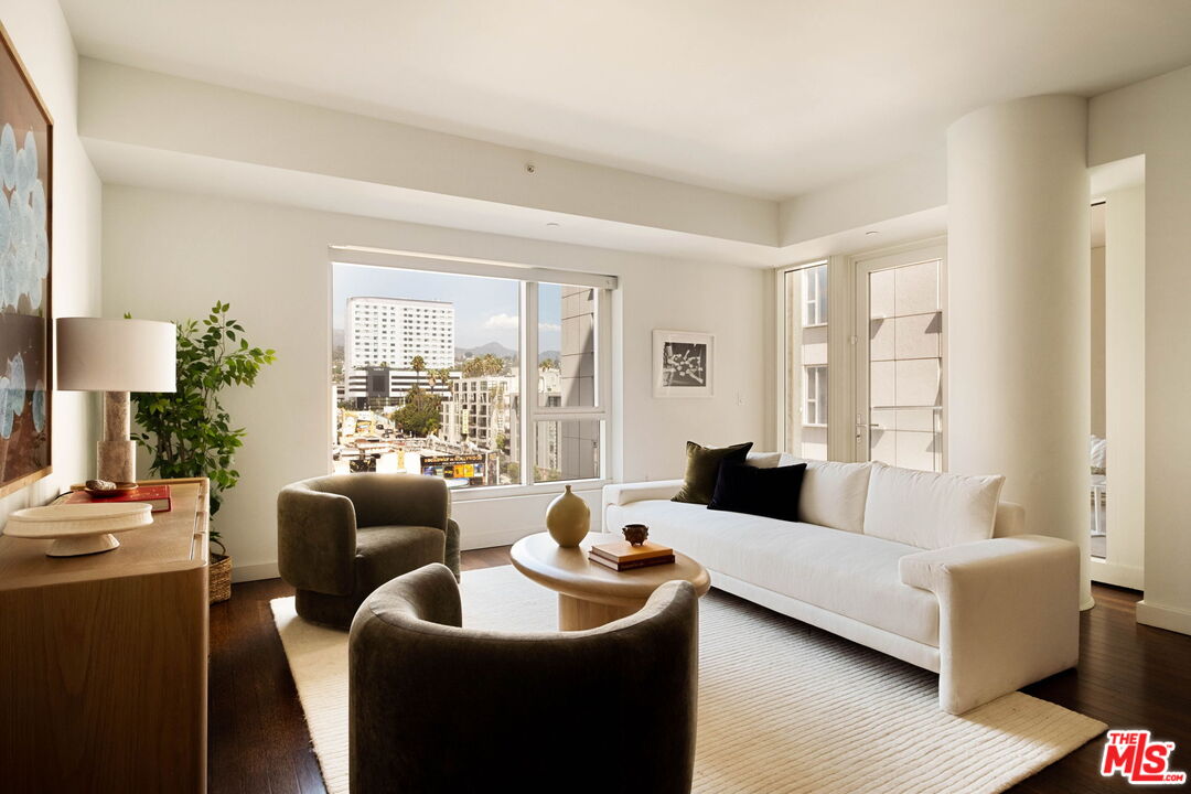 Bright living room with a white sofa, dark armchairs, and large windows overlooking city buildings.