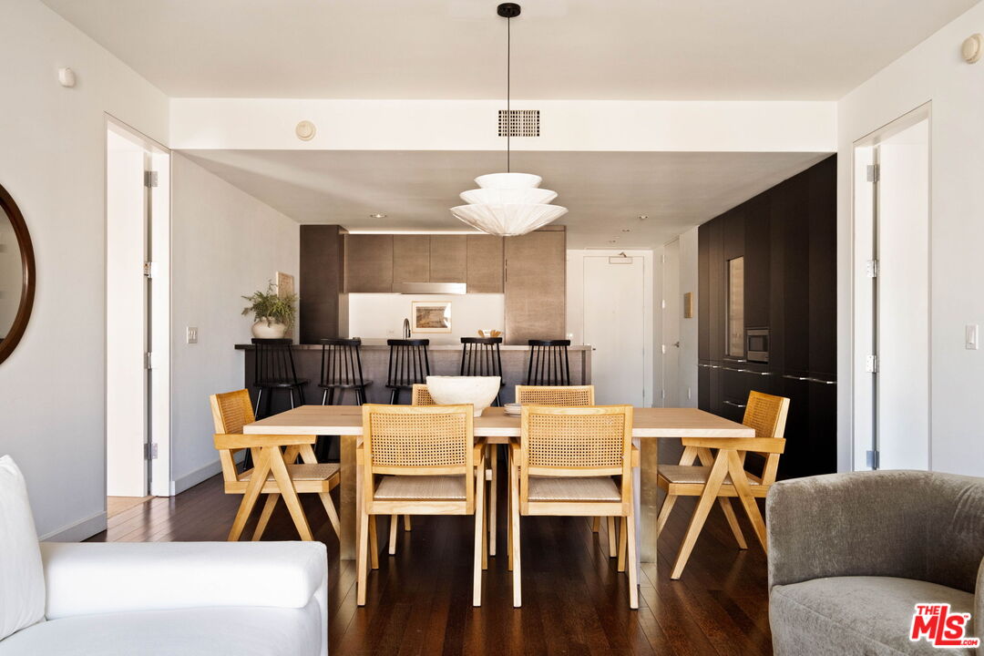 Dining area with a light wood table and woven chairs, opening onto a kitchen island with bar stools.