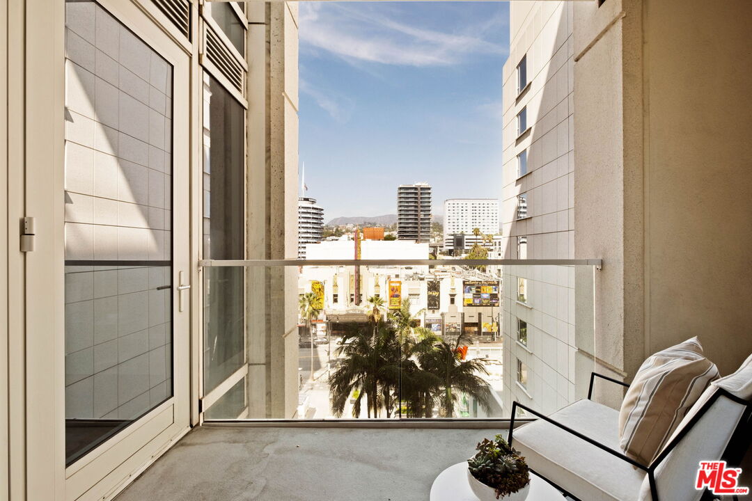 Apartment balcony with glass railing and seating overlooking palm trees and city buildings.