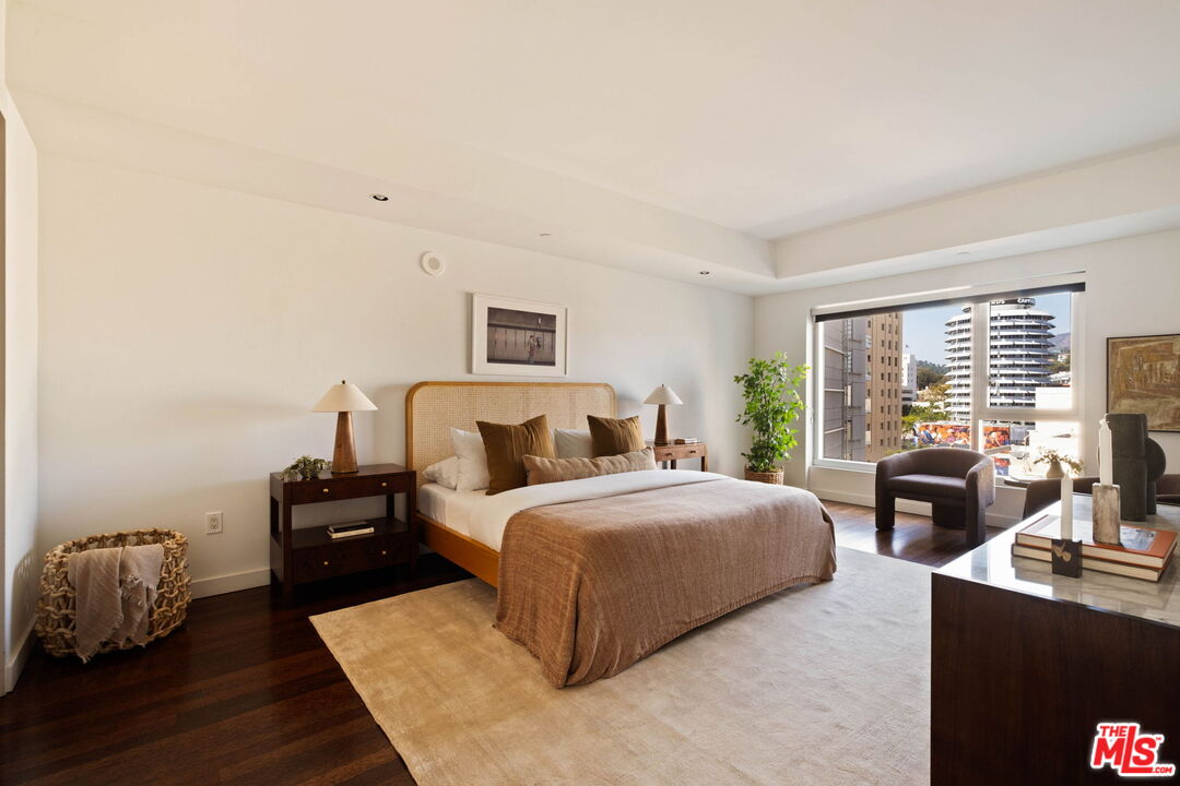 Staged bedroom with a king bed, sitting area, and large windows overlooking city buildings.
