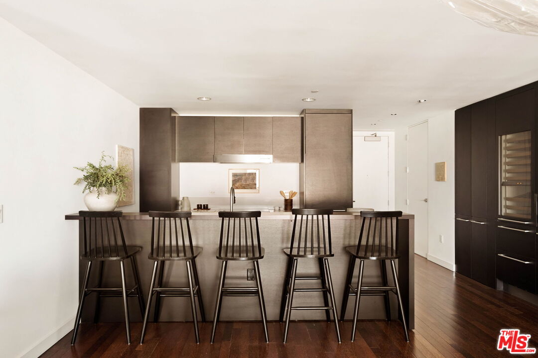 Modern condo kitchen with a long island, five dark bar stools, taupe cabinets, and dark wood flooring.
