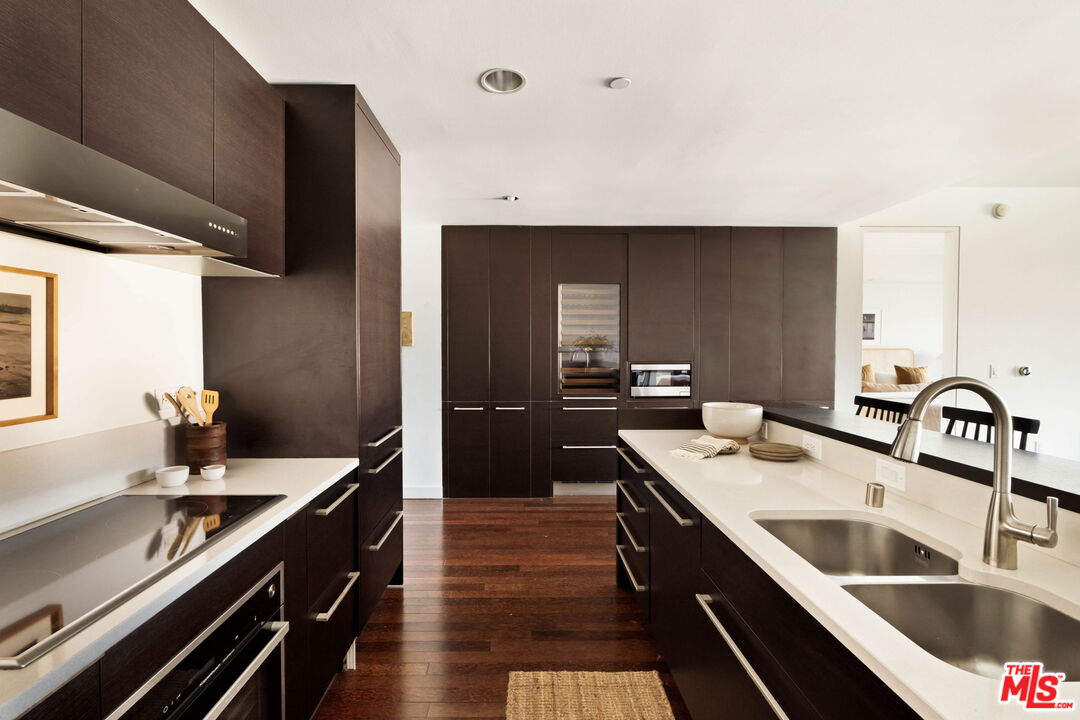 Modern kitchen with dark wood cabinets, white counters, stainless steel appliances, and a large island sink.