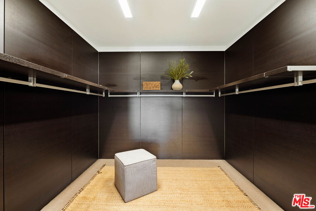 Walk-in closet with dark wood cabinetry, wraparound shelves, a tan rug, and an upholstered stool.