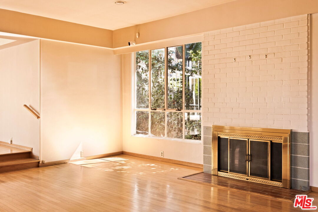 Empty living room with hardwood floors, a brass-trimmed brick fireplace, and a large window.